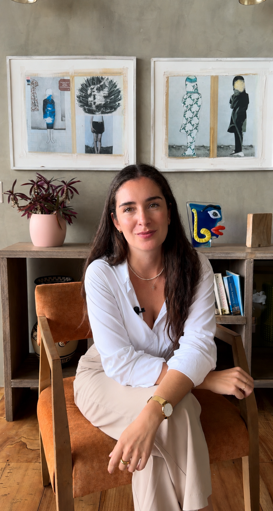 A woman sitting in a brown chair in front of a bookshelf with a pink plant pot, colorful decorative object, and books. Artworks on the wall behind her feature stylized human figures with abstract faces.