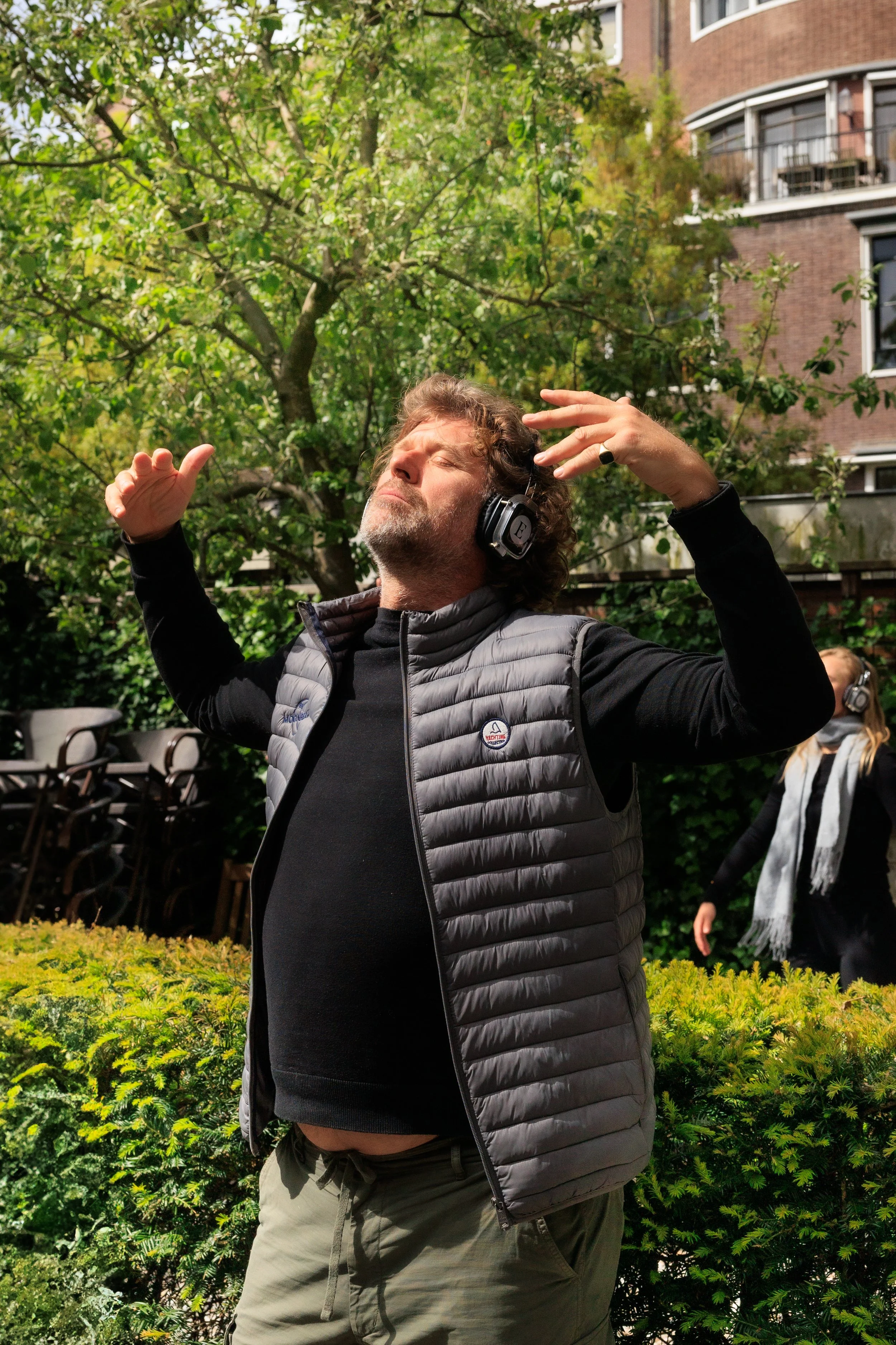 A man with curly hair and a beard is wearing headphones and a black jacket with a gray vest. He is outdoors in a garden with lush green bushes and trees, with a brick building in the background. His eyes are closed, and he appears to be enjoying music with his arms raised.