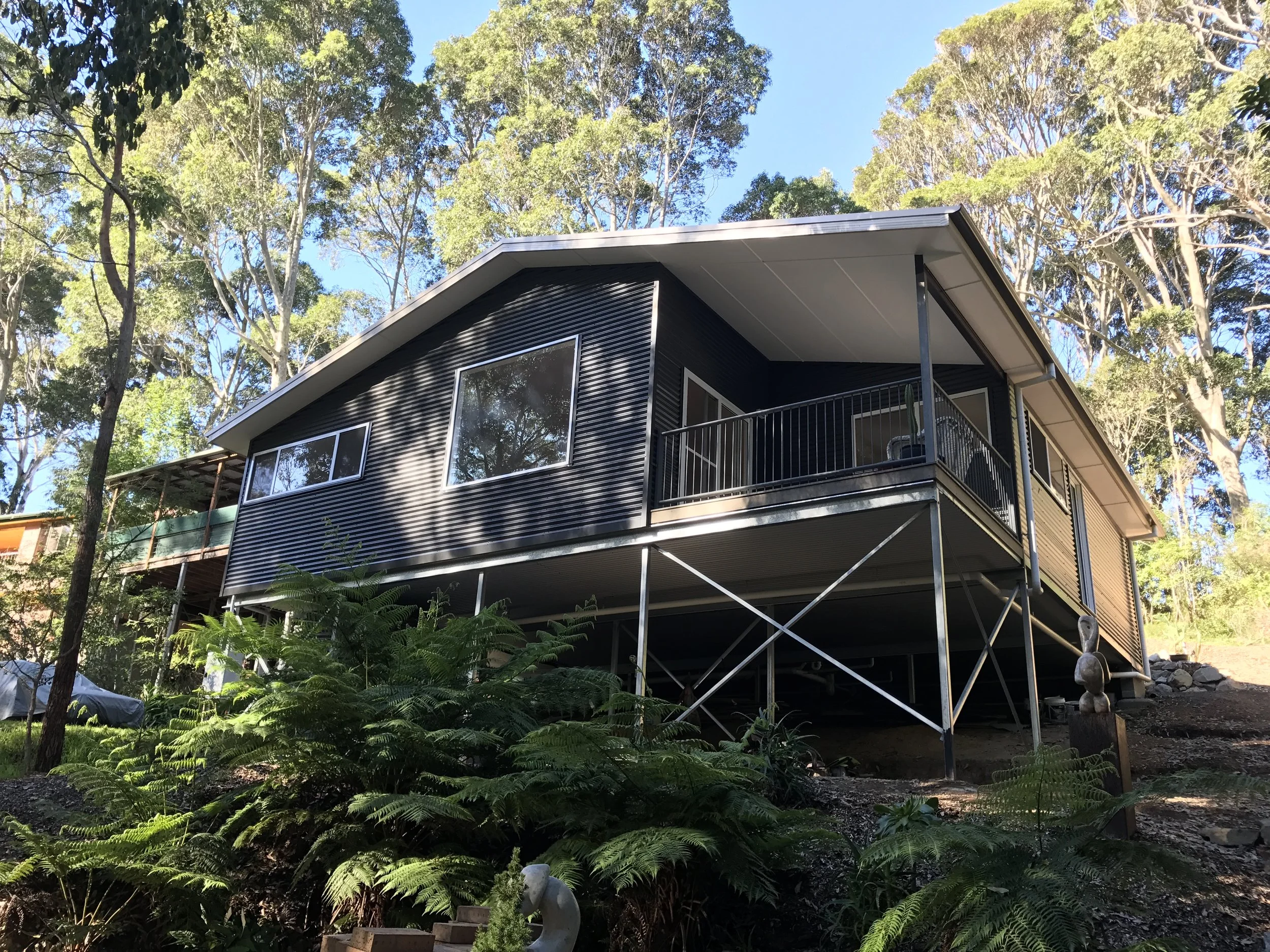 Modern two-story house with black exterior walls and large windows, surrounded by tall trees and lush green foliage, under a clear blue sky.