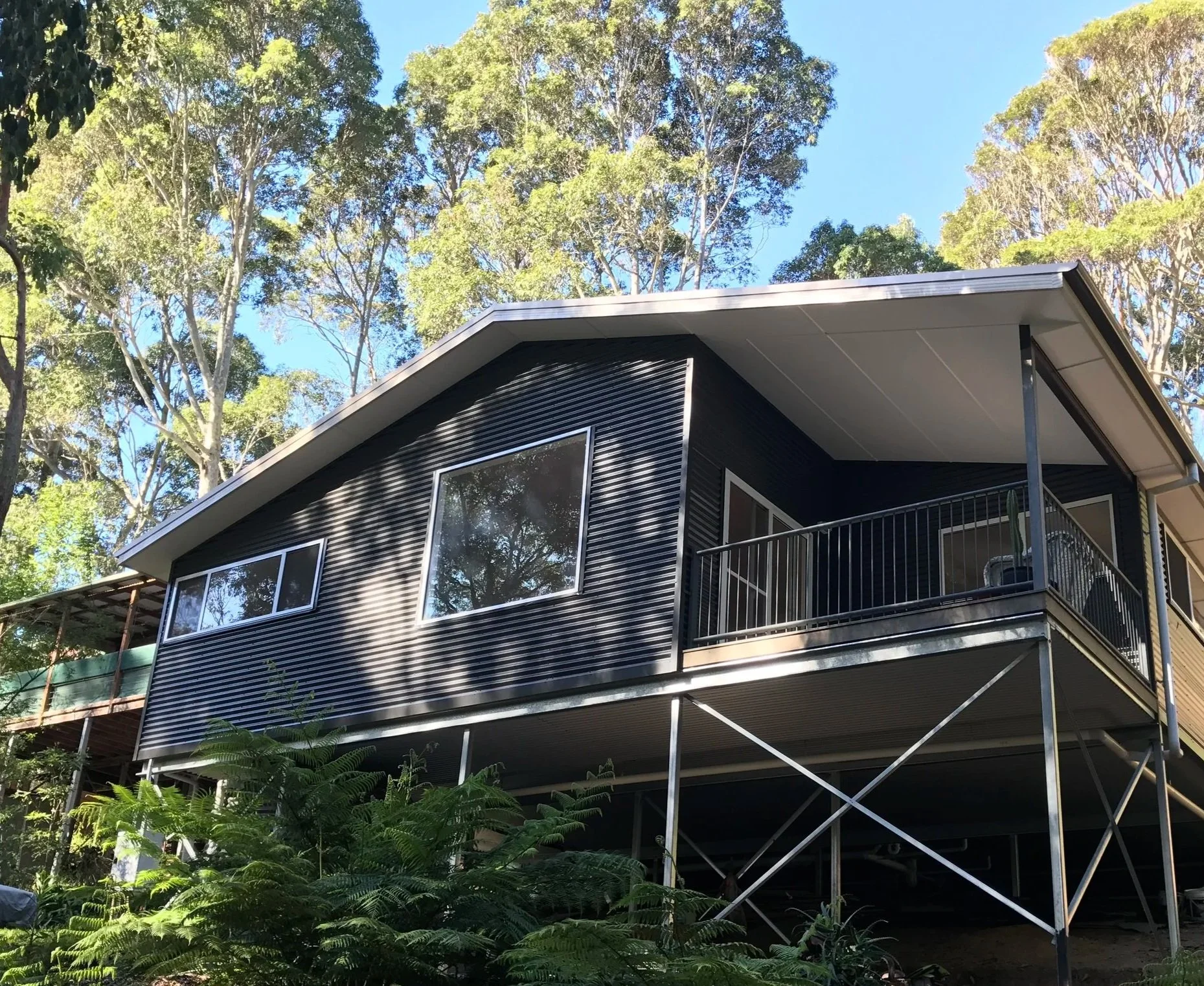 Modern two-story house with black exterior siding, large windows, and a balcony with a metal railing, surrounded by tall trees and green foliage.