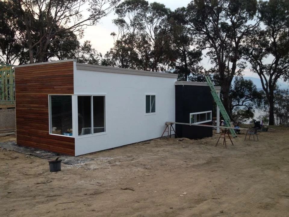 Under construction modern house with white and black exterior, large glass window, and wooden paneling on the side, set in a dirt yard with trees in the background.