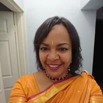 A woman with shoulder-length black hair, wearing a yellow traditional outfit with jewelry, smiling indoors.