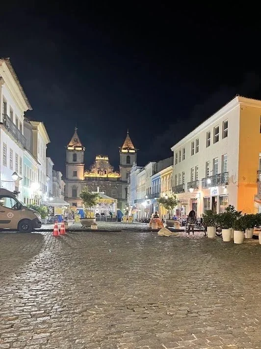 Nighttime view of a cobblestone street in a historic district with colorful buildings, some with outdoor seating, and a church with twin towers illuminated in the background.