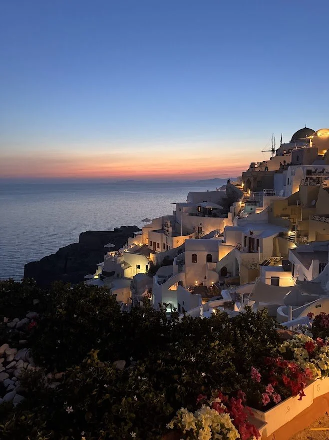 View of white hillside buildings at sunset in Santorini, Greece, overlooking the sea with a colorful sky.