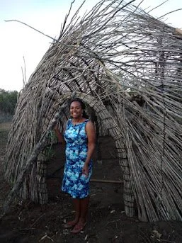 Woman standing in front of a large, rustic, dome-shaped hut made of dried branches.