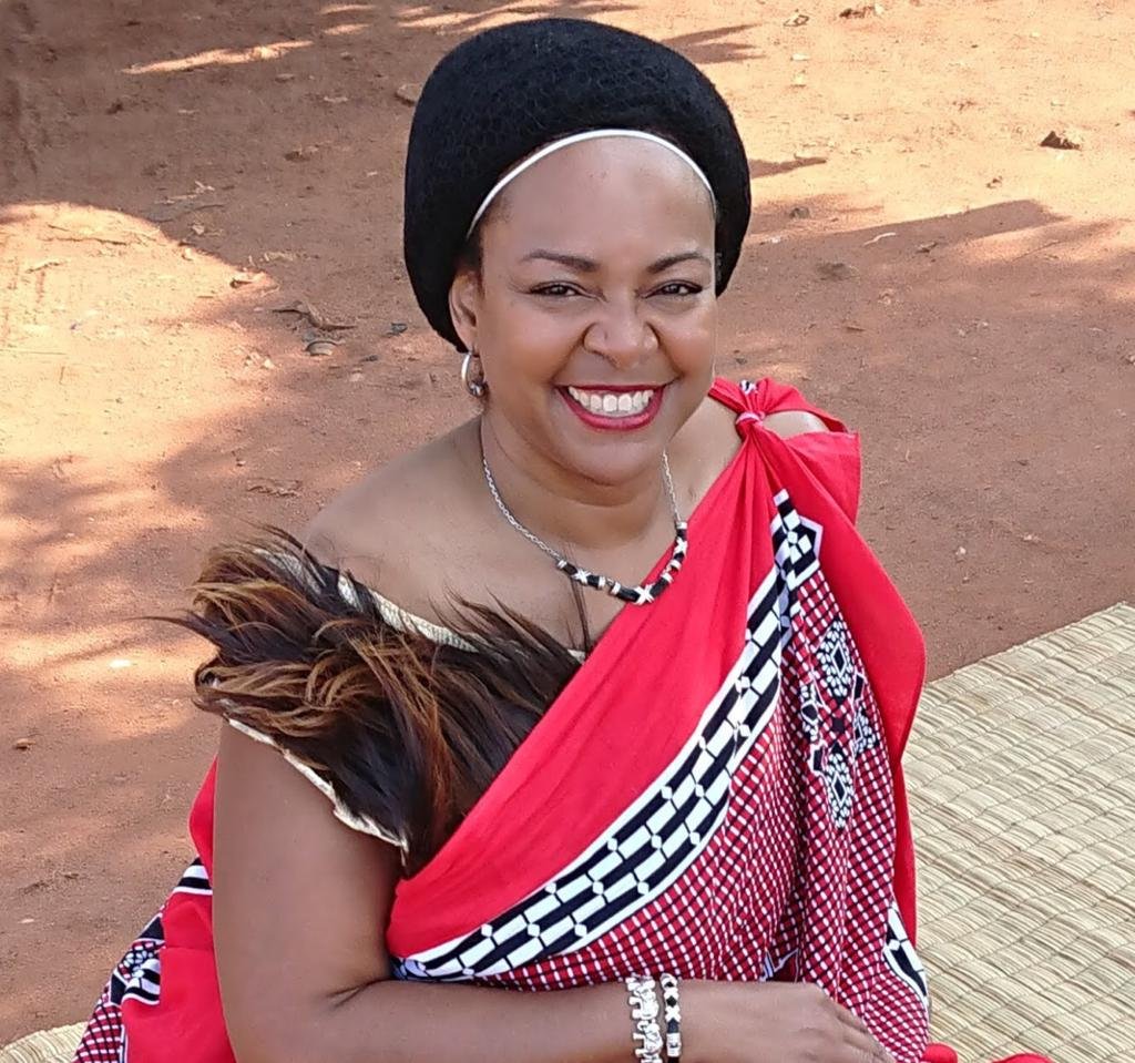 Smiling woman dressed in traditional African attire sitting on a mat outdoors on red dirt ground.