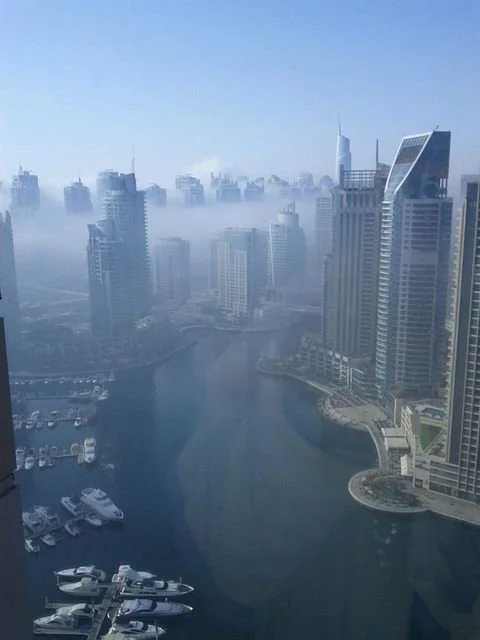 Dubai Marina skyline with tall modern buildings, a marina, and boats docked at the waterfront, shrouded in fog with clear blue sky.