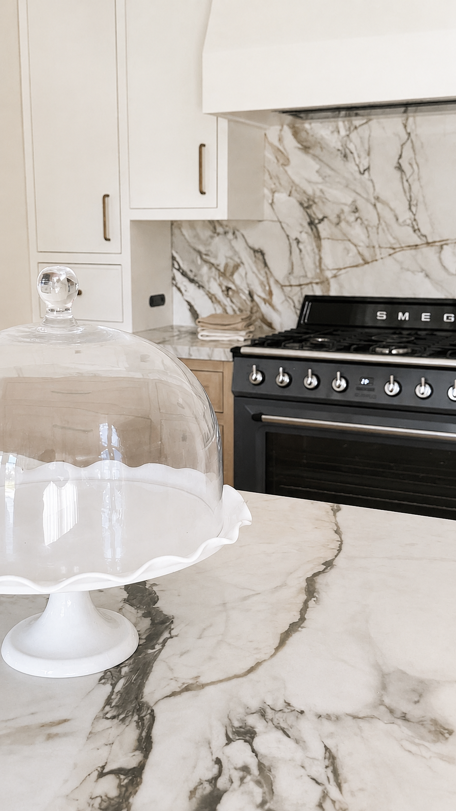 Kitchen with white cabinets, black Smeg stove, marble backsplash, and a marble countertop with a glass cake stand.