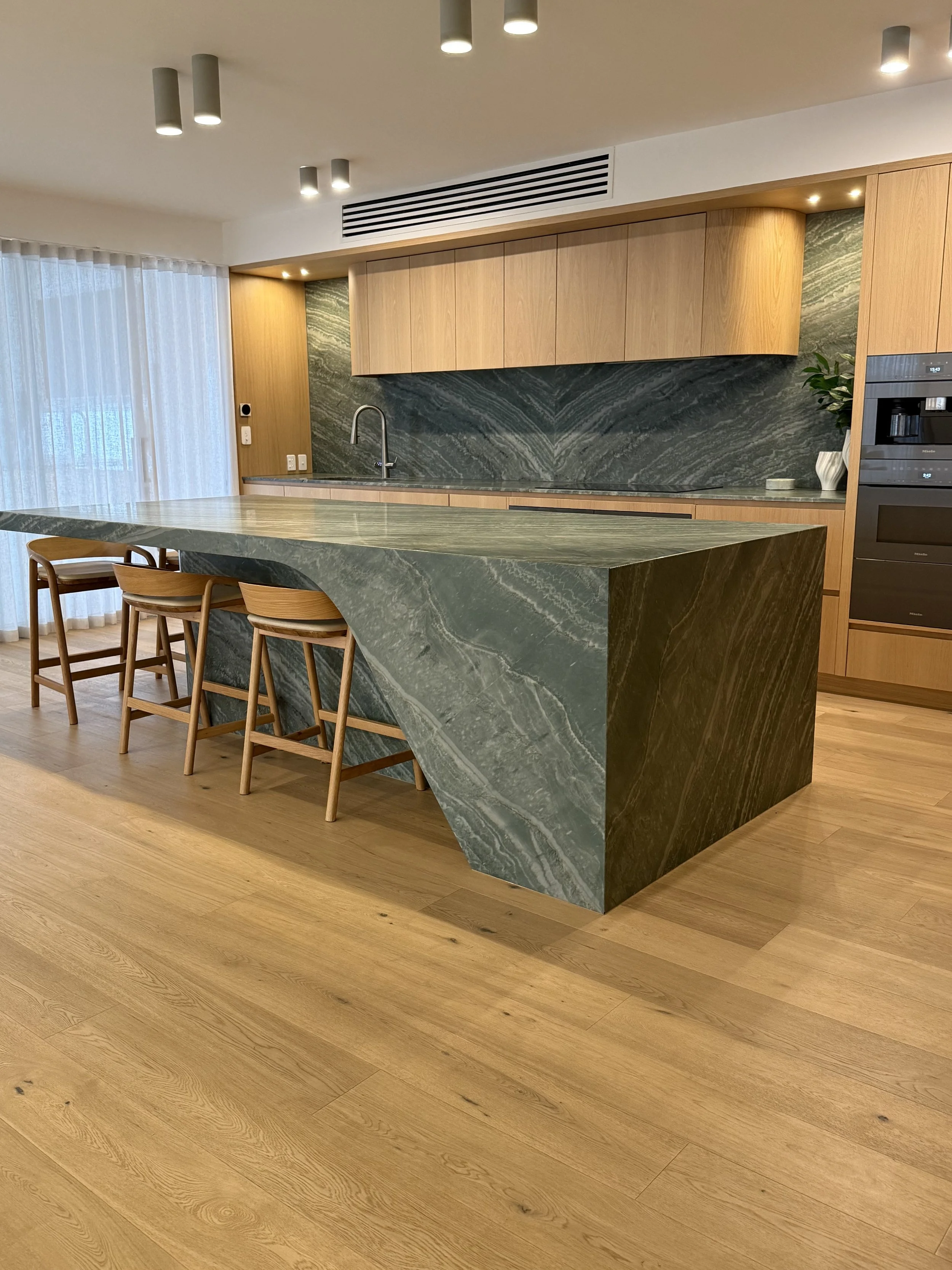 Modern kitchen with green marble island and backsplash, wooden cabinets, wooden chairs, and hardwood flooring.
