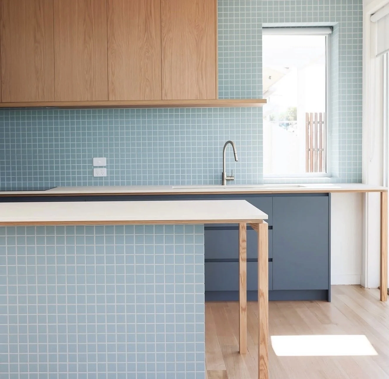 Modern kitchen with blue tile backsplash, wooden cabinets, and a large window letting in natural light.
