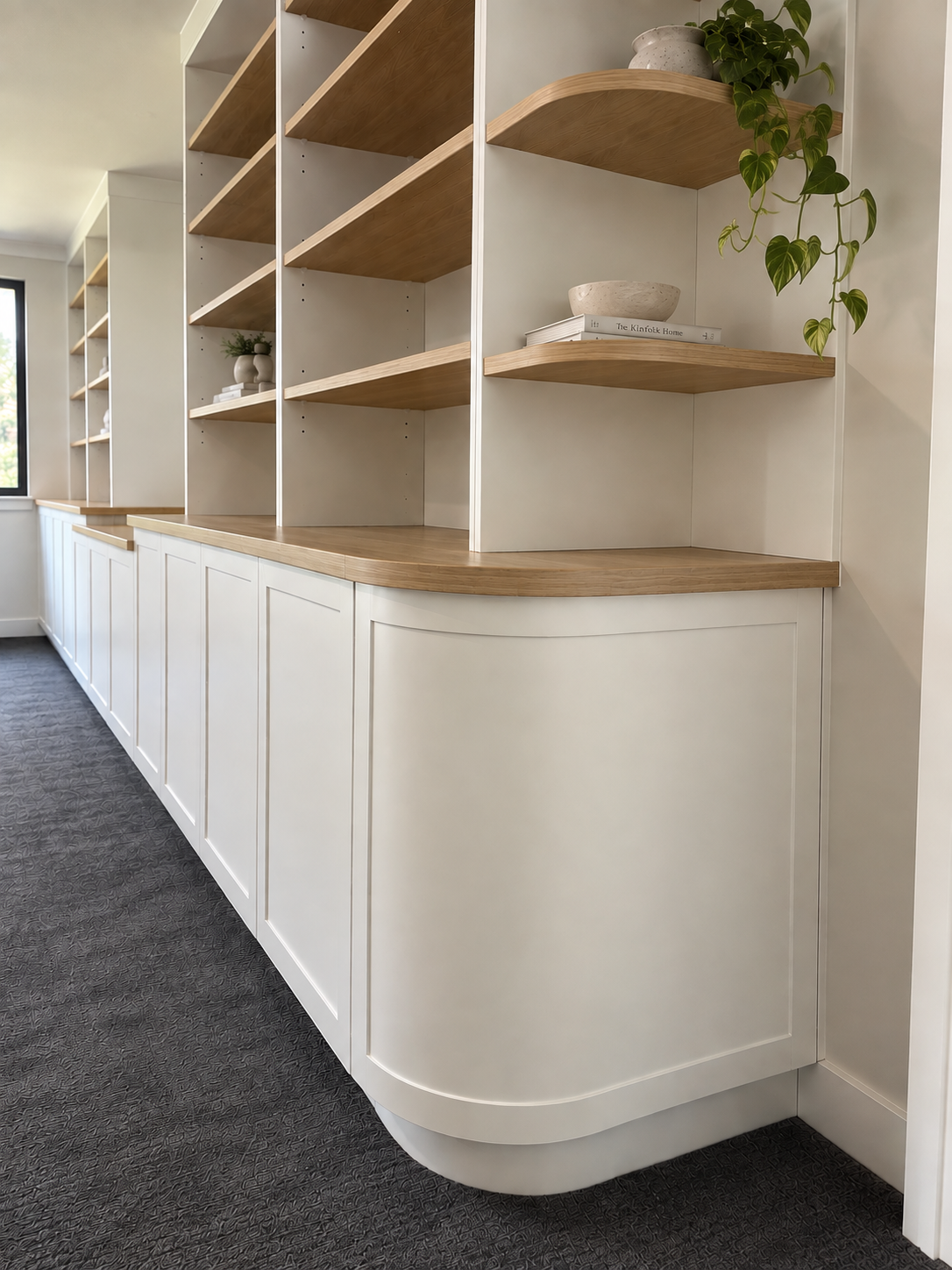 White built-in shelving unit with wooden shelves and cabinet doors, decorated with plants and books, in a room with a window and dark carpet.