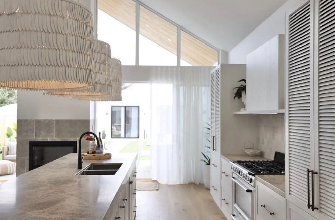 Modern kitchen with white cabinets, a countertop island with a sink, stove, and a large woven lampshade hanging above the island, natural light coming through large windows with white curtains.