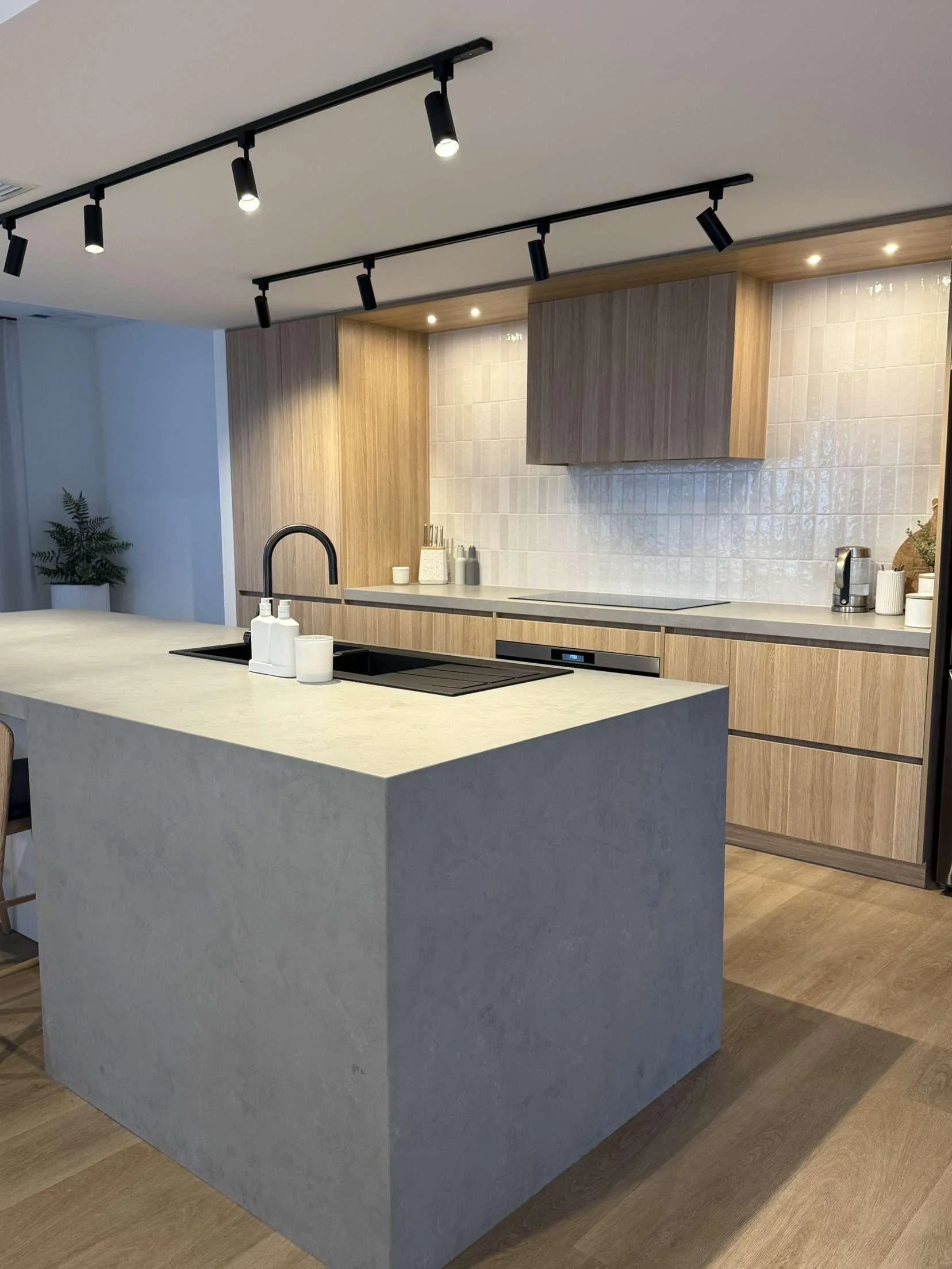Modern kitchen with a large light-colored island, black sink, wooden cabinets, and track lighting on the ceiling.