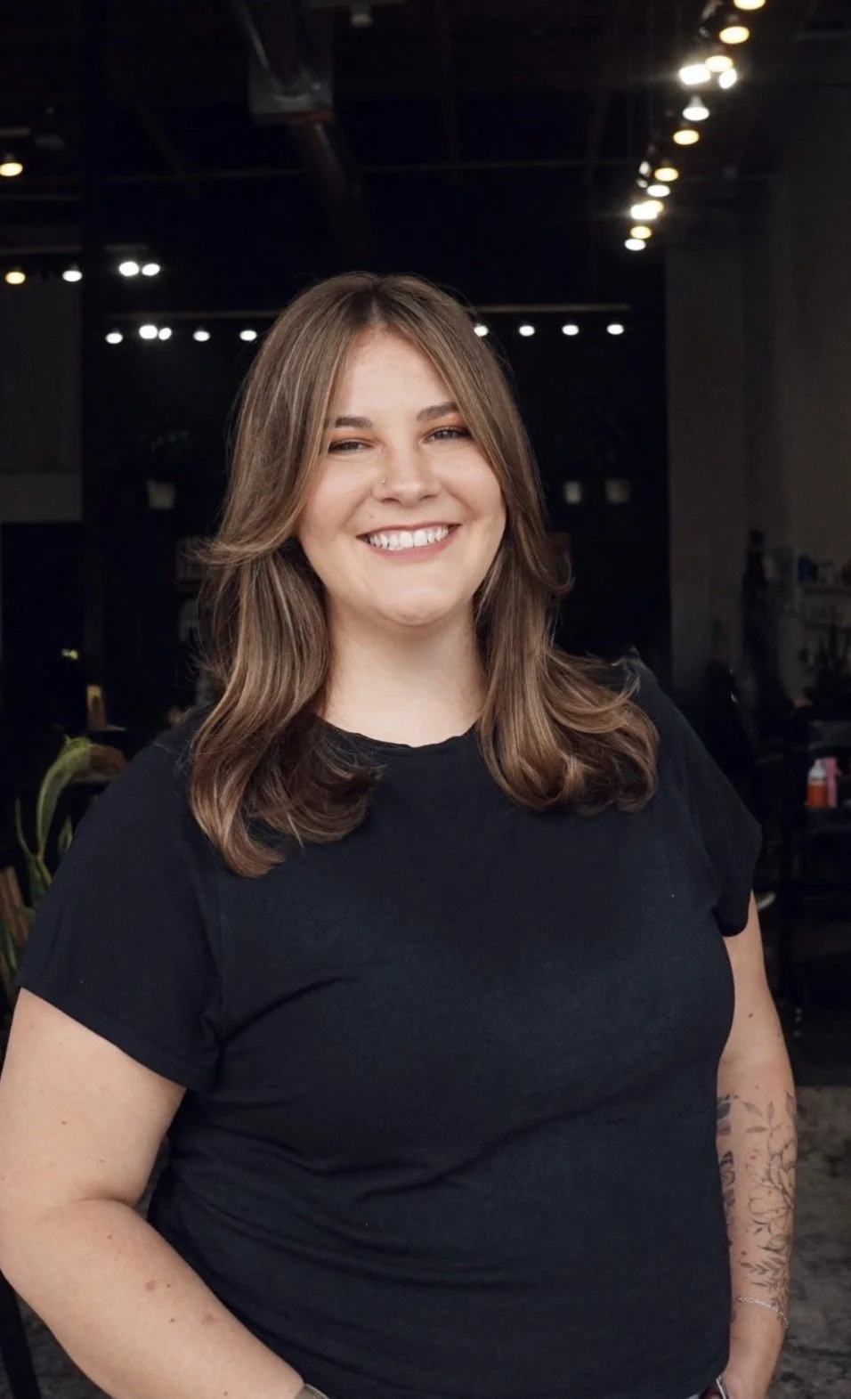 A smiling woman with shoulder-length wavy brown hair, wearing a black T-shirt, standing indoors with dim lighting and blurred background.