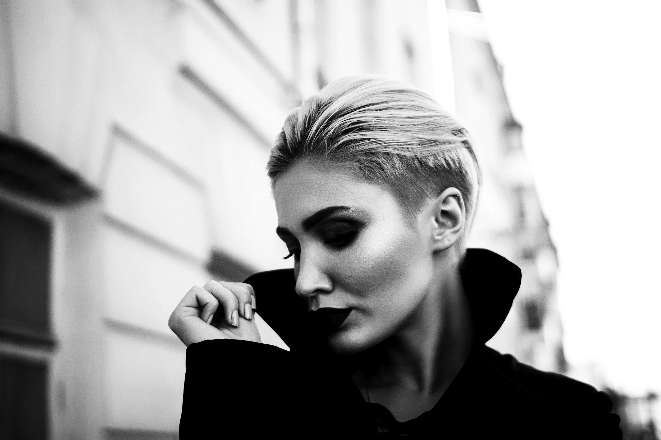 Black and white close-up photo of a woman with short, styled hair, looking downward, with her hand near her face, set against a blurred urban background.