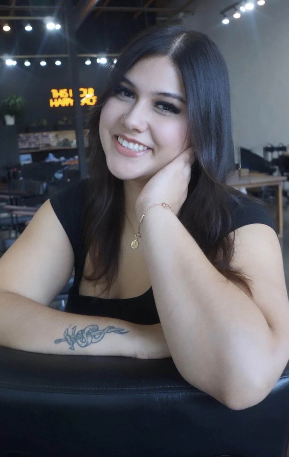 A woman with dark hair and makeup sitting at a table in a cafe, smiling, with a tattoo on her forearm and jewelry on her wrist and neck, in a modern cafe setting with warm lighting.