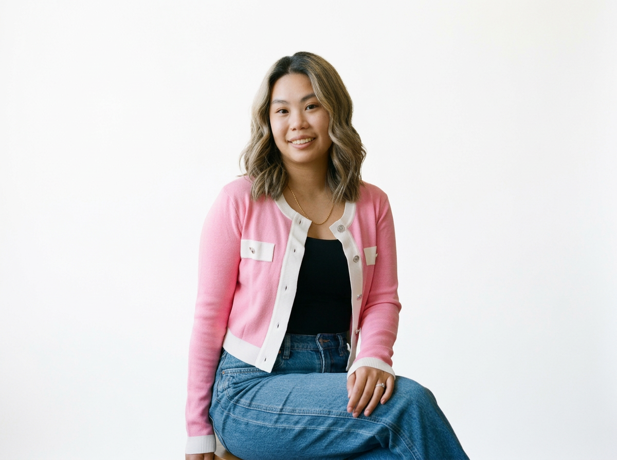 A young woman with shoulder-length wavy hair, wearing a pink cardigan over a black top, sitting with one knee bent and smiling against a plain white background.