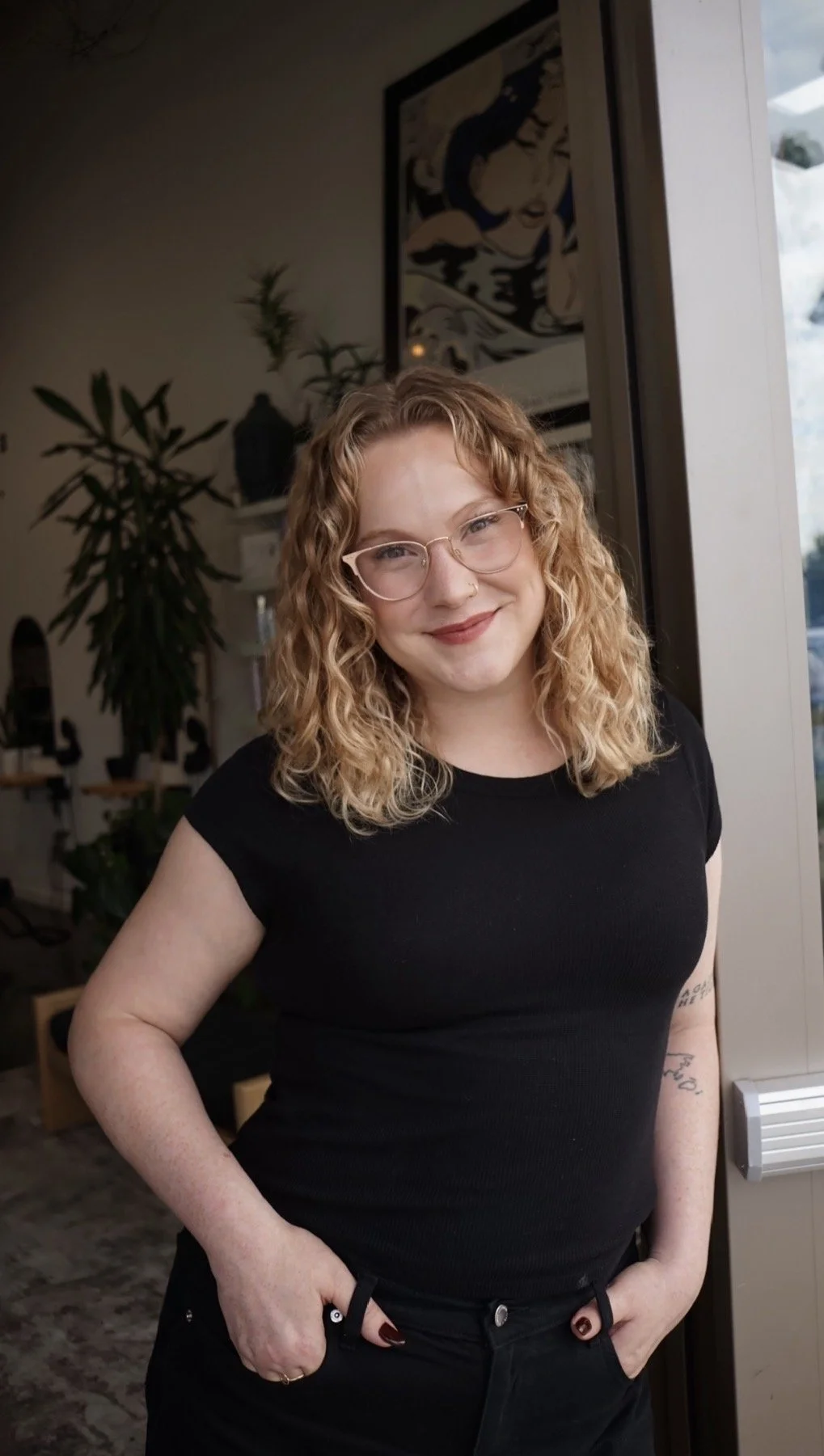 A woman with curly red hair, glasses, and a nose ring smiling while standing indoors near a window, with a plant, artwork, and furniture in the background.