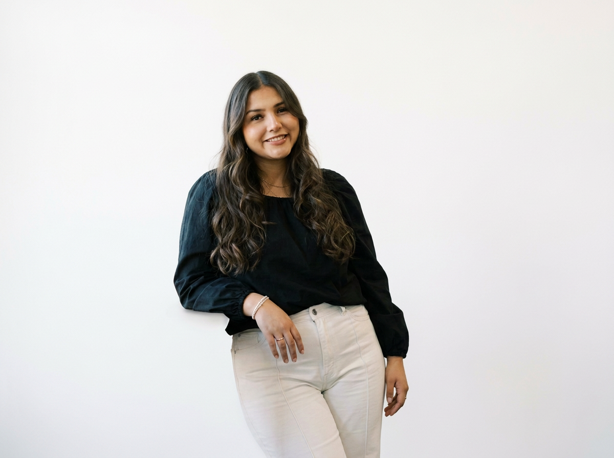 A young woman with long wavy brown hair standing against a plain white background, smiling and wearing a black long-sleeved blouse and beige pants.