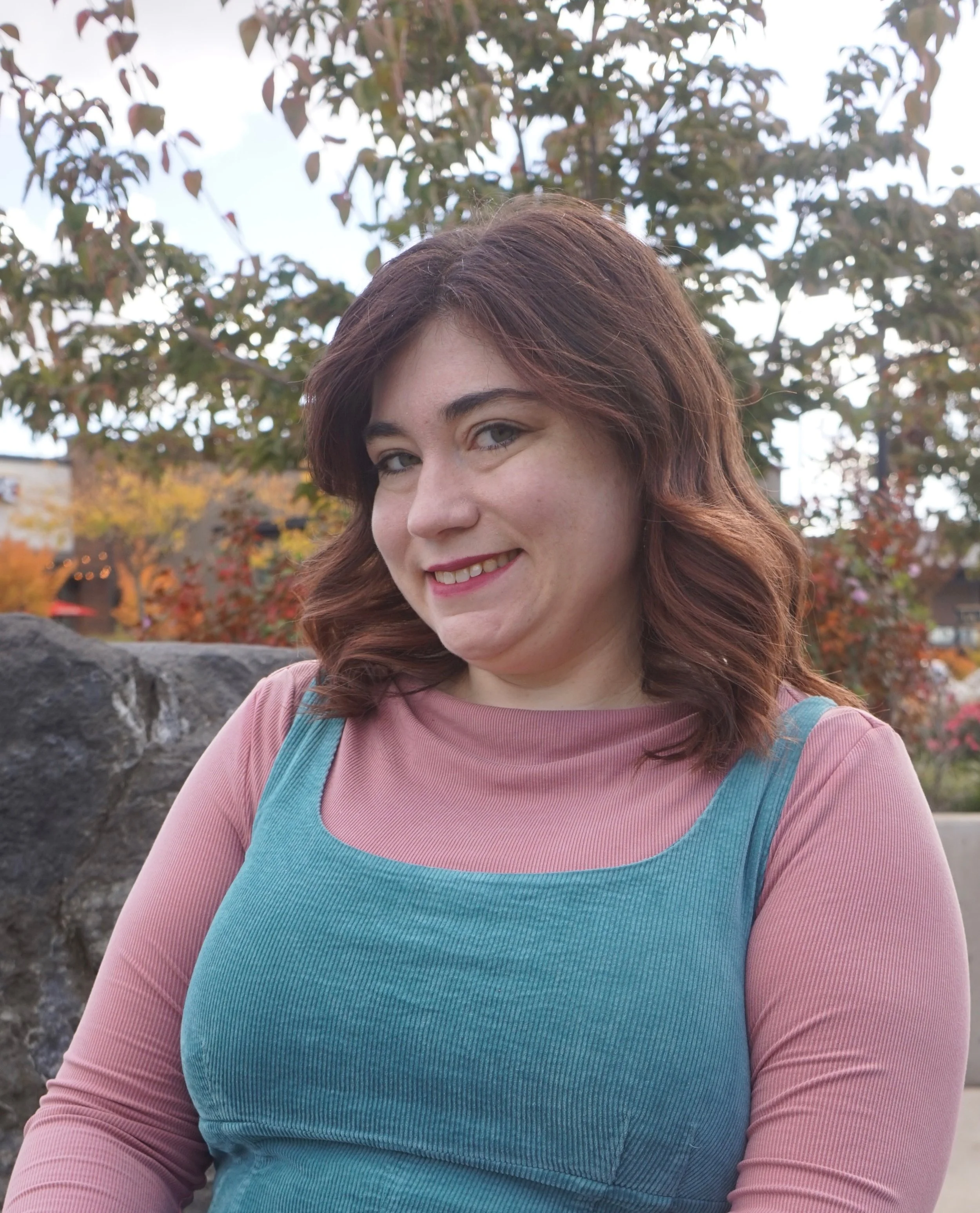 A woman with reddish-brown hair smiling outdoors, wearing a pink and teal long-sleeve shirt, with trees and a cloudy sky in the background.