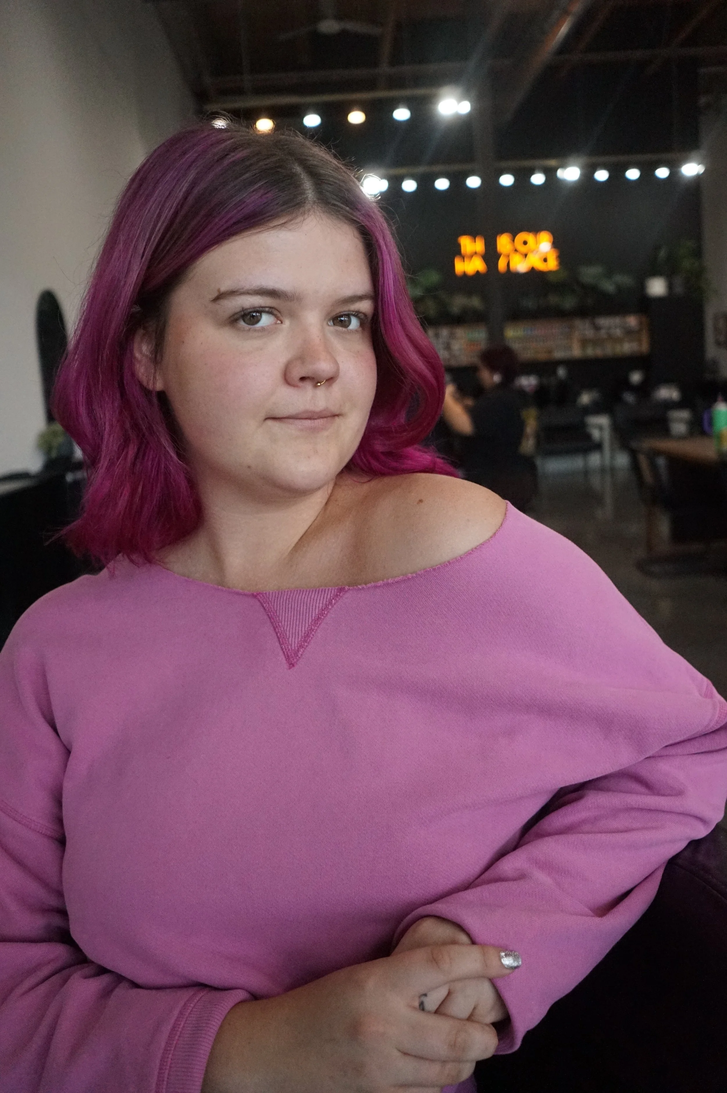 A young woman with shoulder-length purple hair and a septum piercing, wearing a pink off-the-shoulder top, sitting in a dimly lit restaurant or cafe with a black background and a neon orange sign, and a person with red hair in the background.