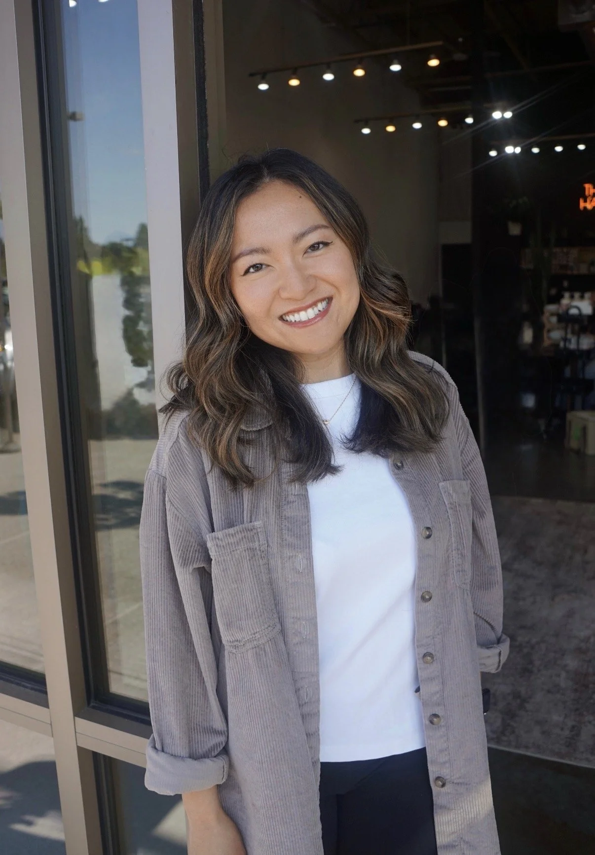 A smiling woman with wavy, shoulder-length hair standing outside a building, wearing a light brown button-up shirt over a white T-shirt.