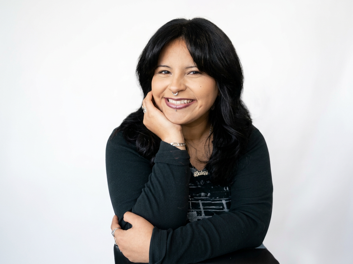 A woman with black hair, nose piercing, and a smile, posing against a plain white background.