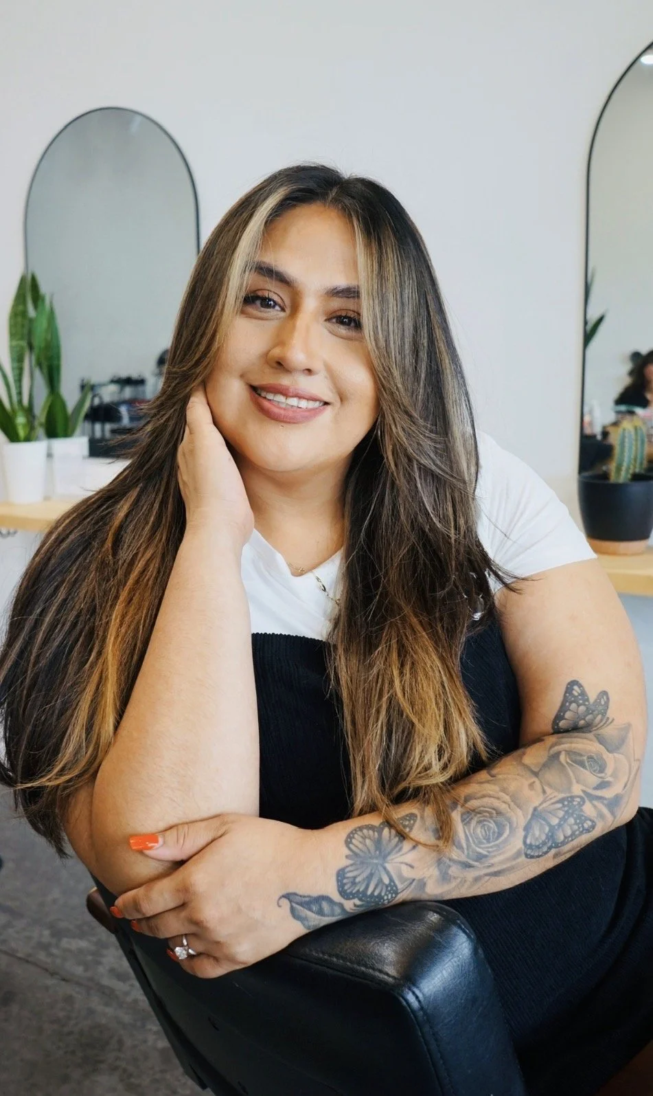 A woman with long brown hair and tattoos on her arm, smiling and resting her head on her hand, sitting in a salon or similar setting.