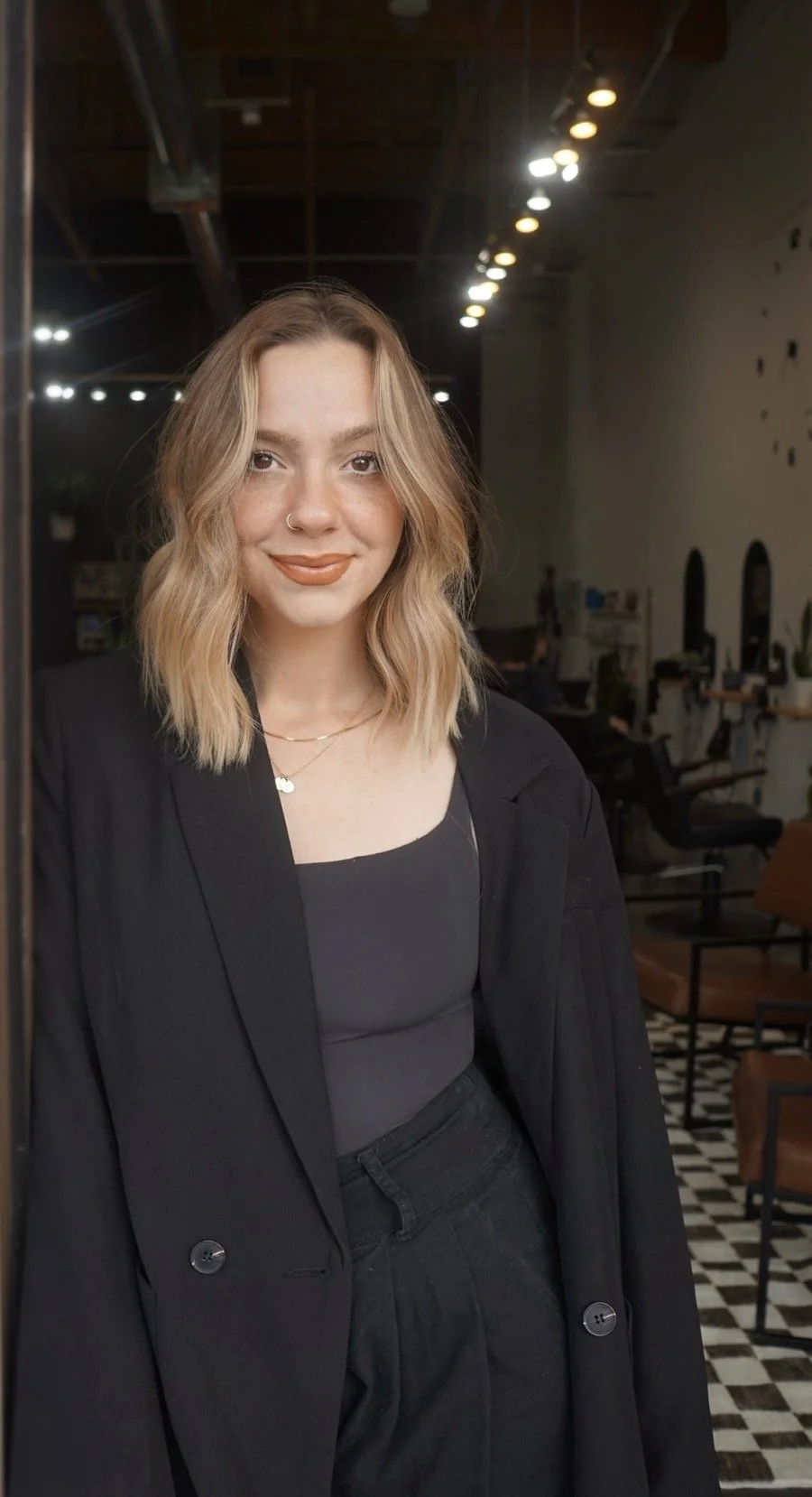 A young woman with shoulder-length wavy blonde hair, wearing a black blazer over a black top, standing inside a stylish cafe with modern decor and seating in the background.