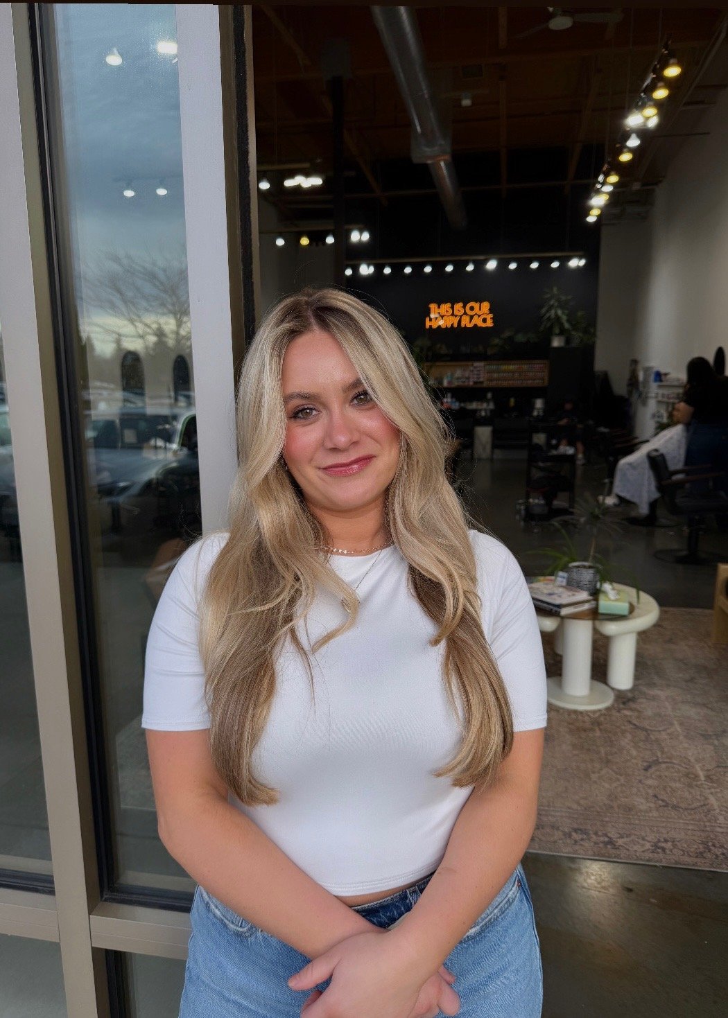 A young woman with long blonde hair, wearing a white t-shirt and blue jeans, is standing inside a coffee shop or cafe near a glass door, smiling at the camera. The background shows the interior of the cafe with some seating and a neon sign.