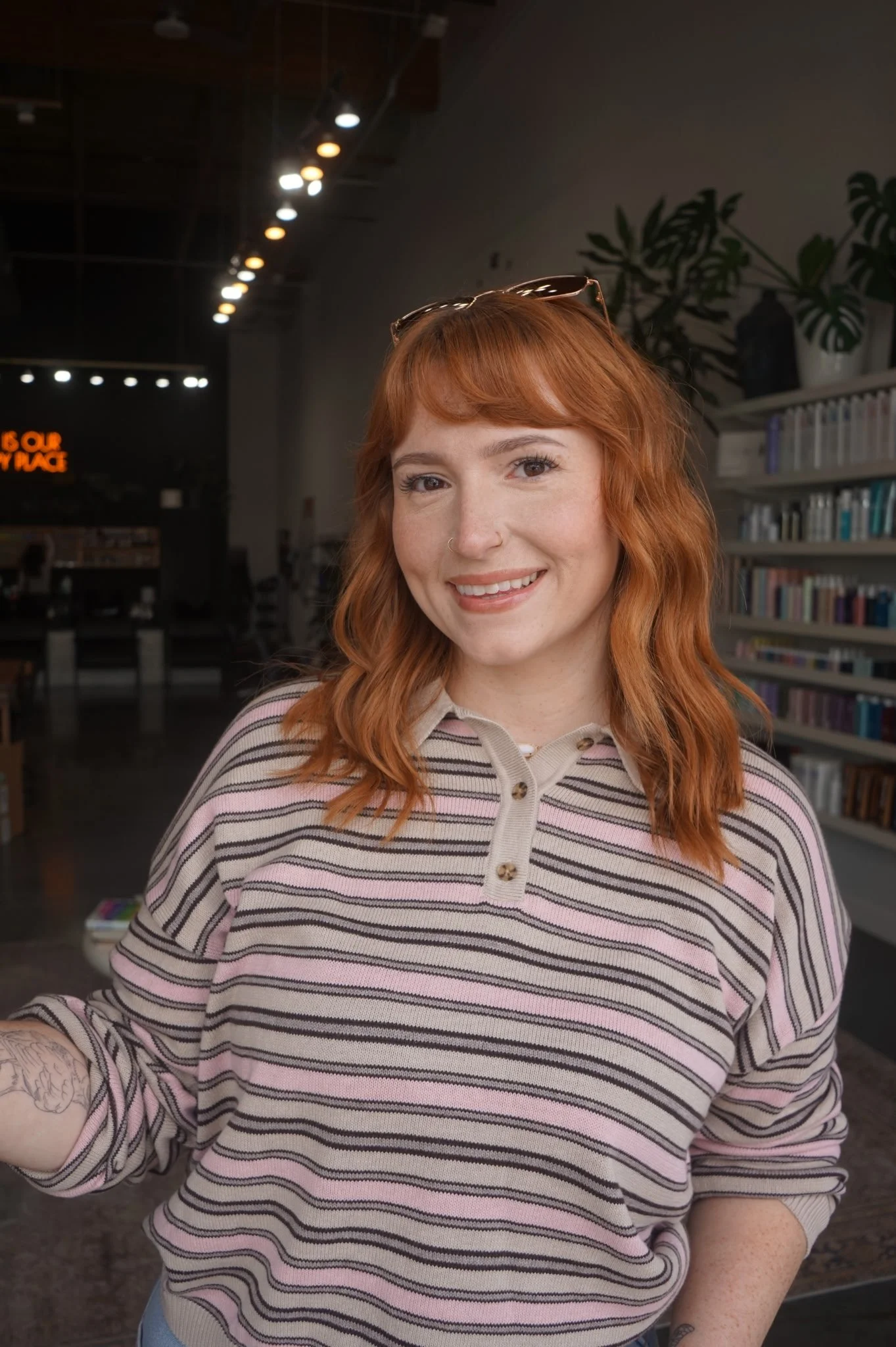 A woman with red hair, glasses on her head, a nose ring, and freckles, smiling in a room with bookshelves and plants in the background, wearing a striped pink, beige, and brown shirt.