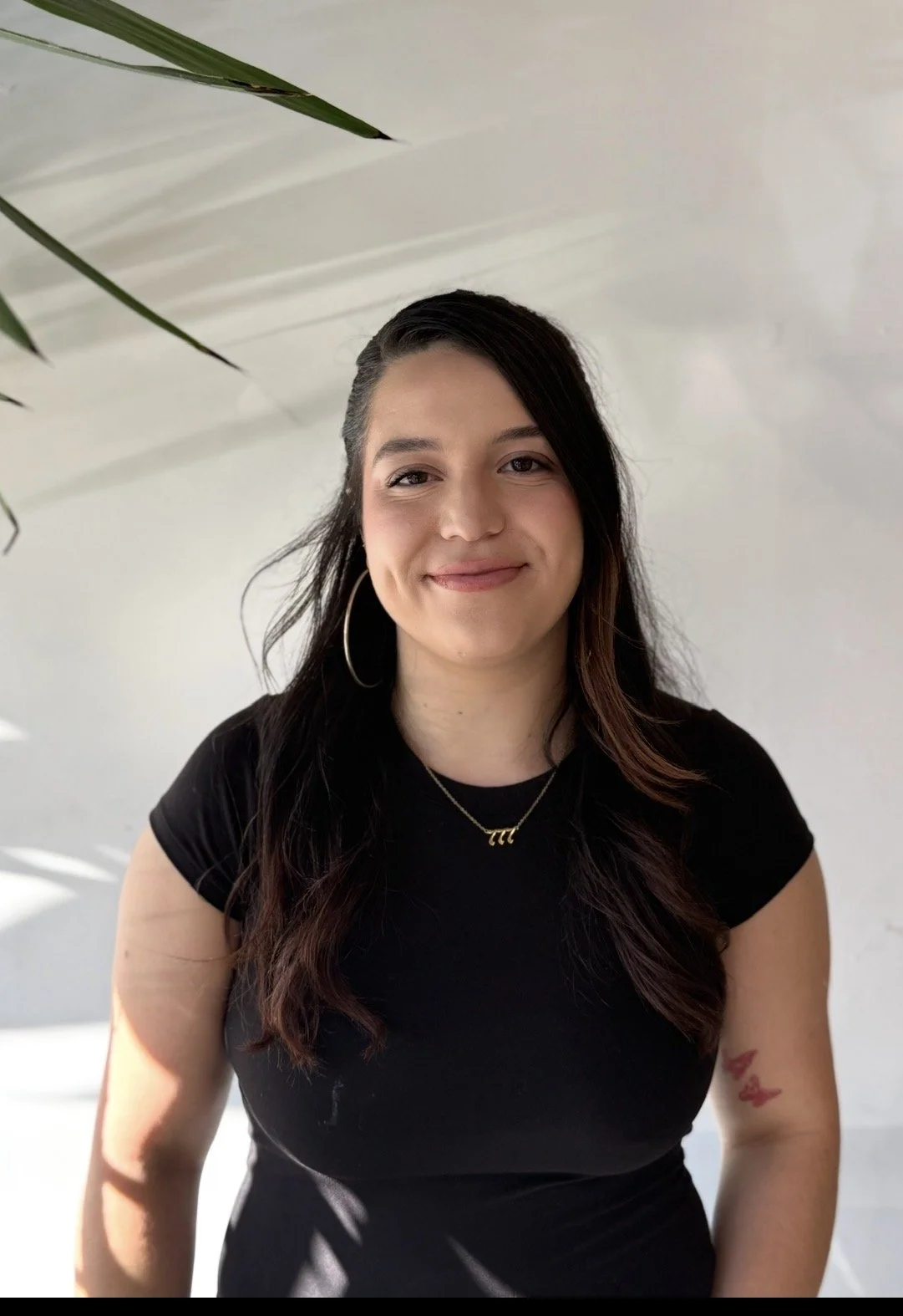 A young woman with long dark hair, wearing a black t-shirt, hoop earrings, and a gold necklace with a letter 'A'. She is smiling and standing in front of a plain light-colored wall.