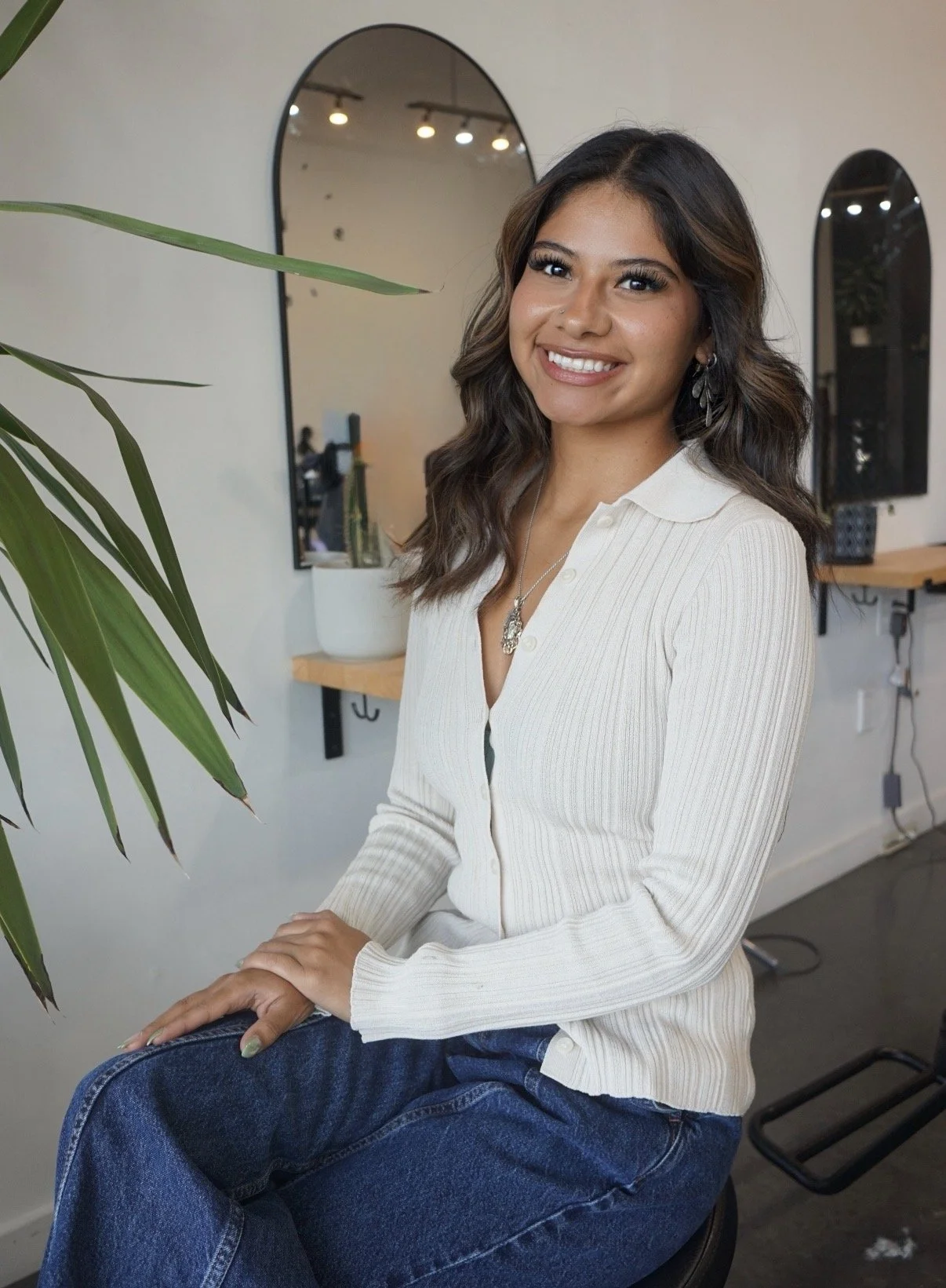 A young woman with long, wavy brown hair and earrings, smiling and sitting in a modern indoor space with mirrors and greenery.