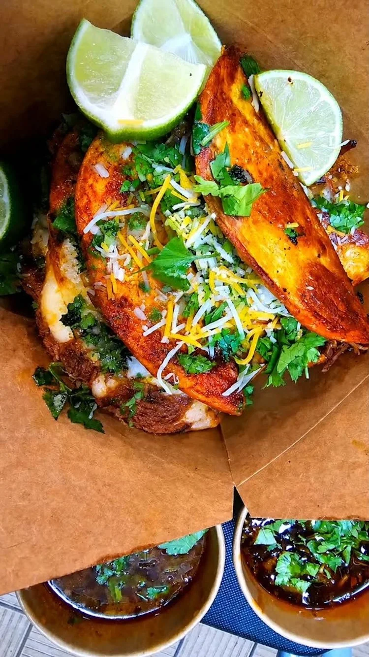 Grilled fish topped with chopped cilantro, shredded cheese, and sliced lime wedges, served on a brown paper-lined tray with bowls of dark sauce on the side.