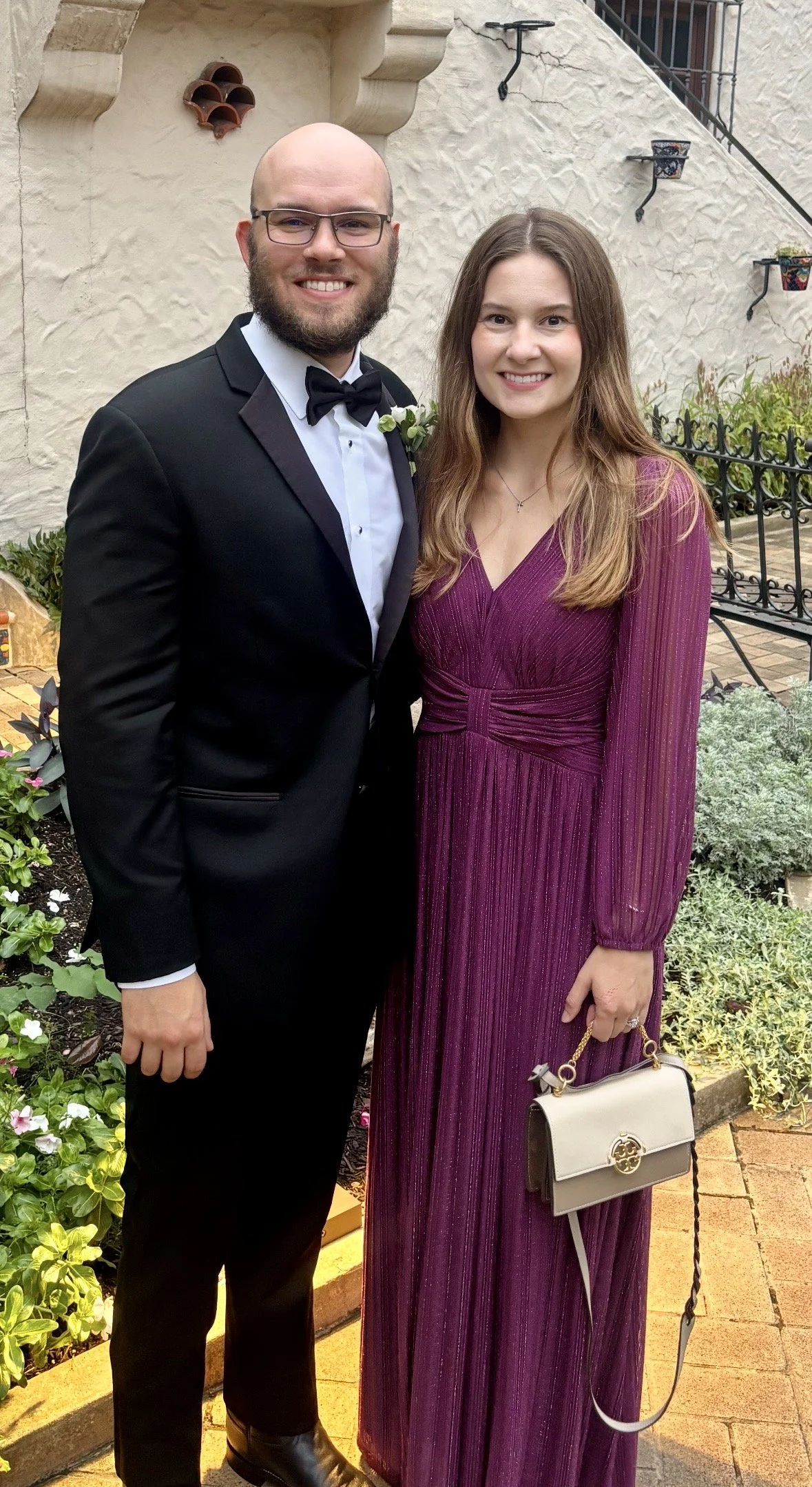A man in a tuxedo and a woman in a purple dress standing side by side outdoors, smiling at the camera.