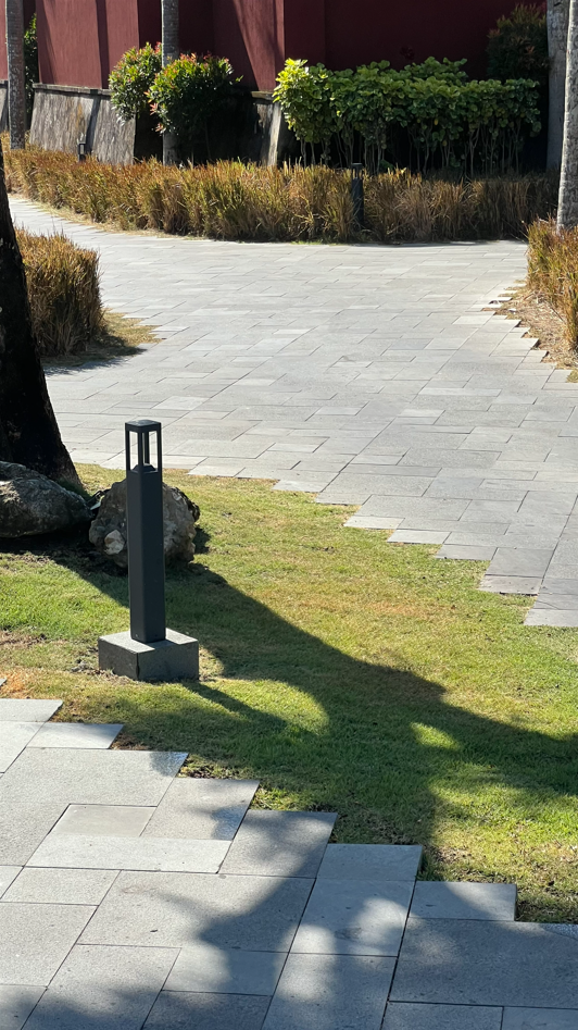 A paved outdoor walkway with a modern post light, bordered by grass, rocks, and bushes, with shadows cast by nearby trees.