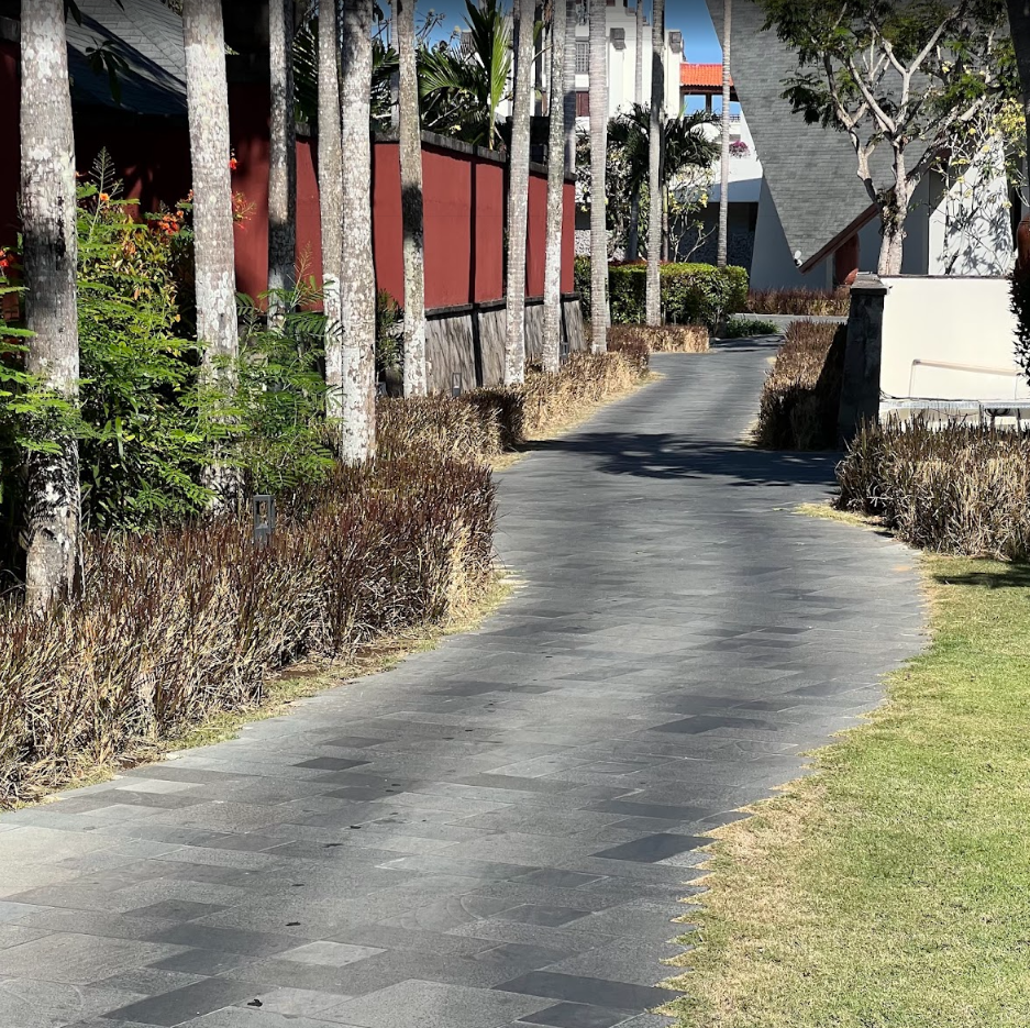 A winding paved walkway flanked by bushes and trees, with a red fence on the left and white buildings in the background, under sunny skies.