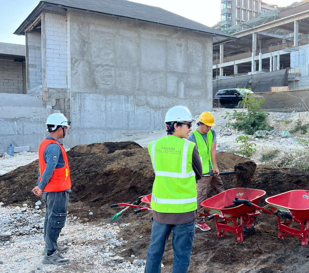 Three construction workers wearing safety helmets and vests working on a site with dirt, wheelbarrows, and a partially built structure in the background.