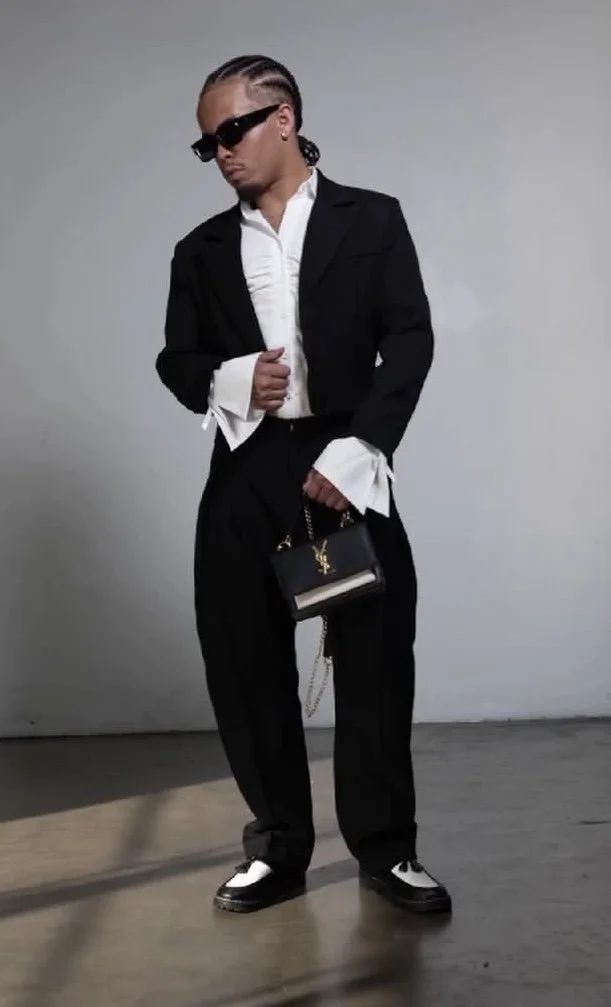 Person wearing black suit, white shirt, sunglasses, holding a designer handbag, standing indoors against a plain wall.