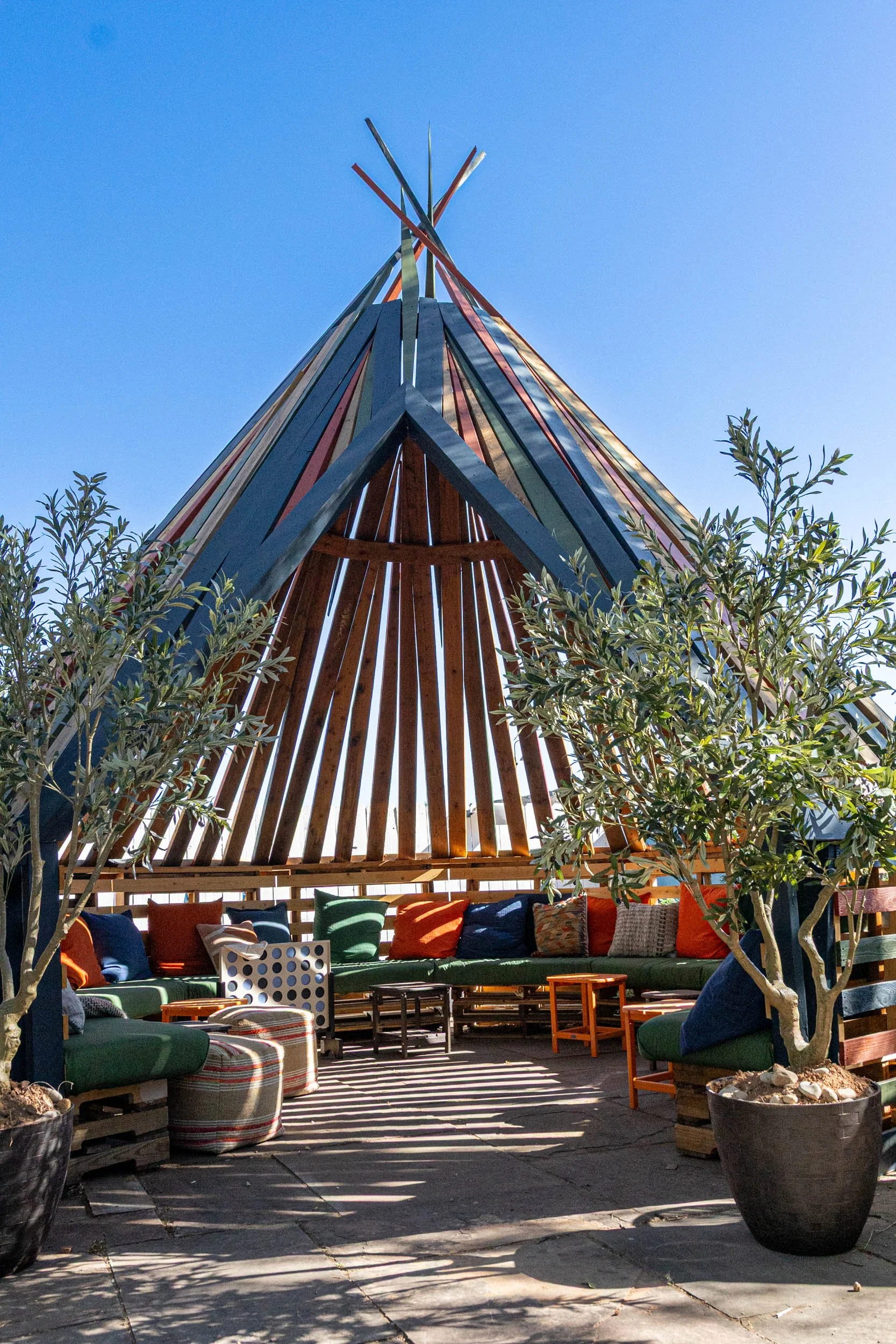 Outdoor seating area with colorful cushions, potted trees, and a modern wooden structure with a tall, pointed roof.