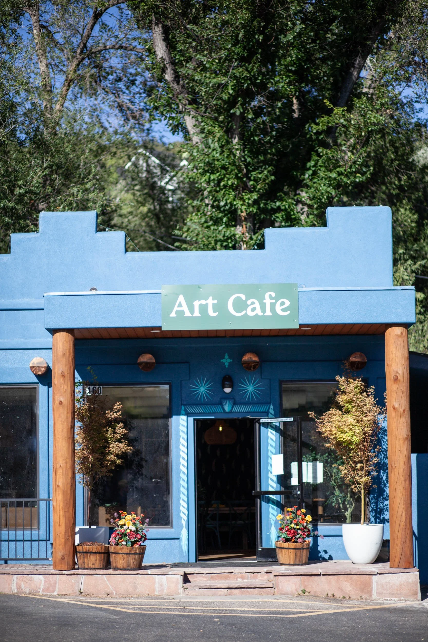 Blue building with a sign that reads "Art Cafe" above the entrance, decorated with potted plants and flowers outside. Large windows showing the interior, with trees and a clear sky in the background.