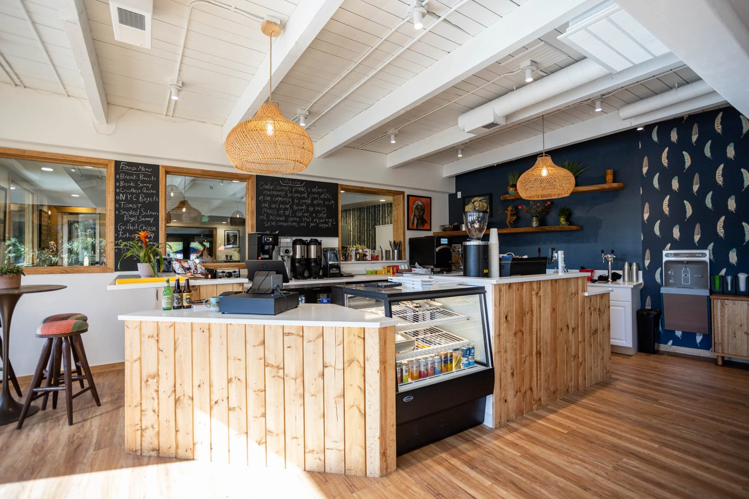 Interior of a modern cafe with wooden accents, a counter with a cash register, a blackboard menu, pendant woven rattan lights, and a blue wall with a feather pattern, decorated with artwork and potted plants.