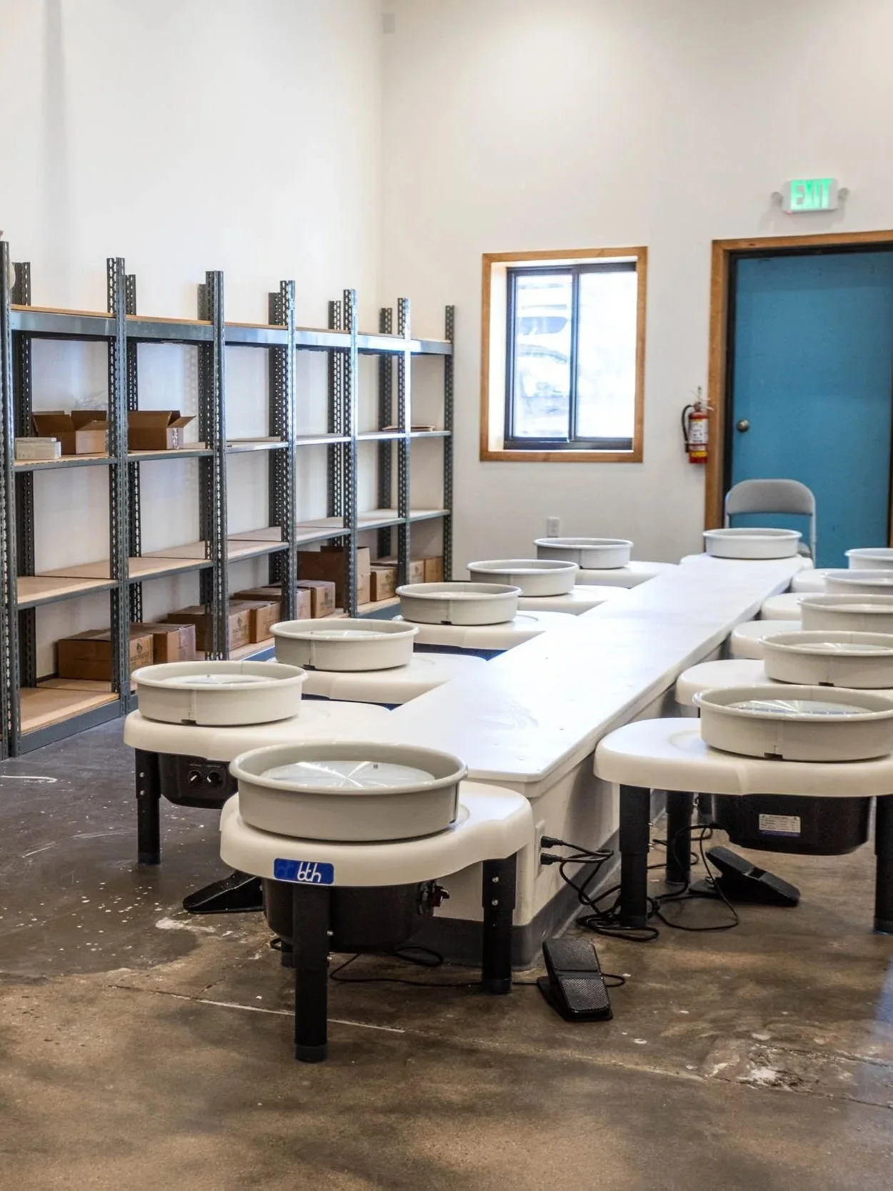 A pottery workshop with multiple pottery wheels and shelves stocked with boxes and pottery supplies, illuminated by natural light from a window.