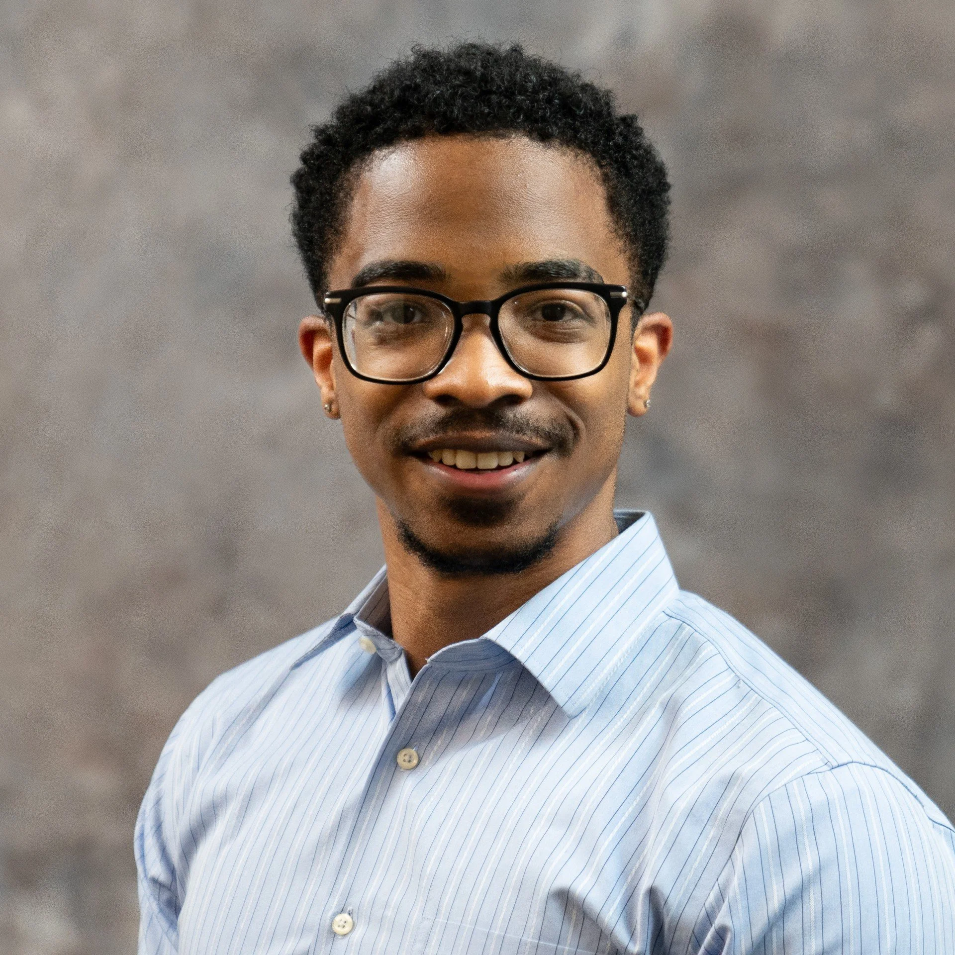 A smiling man with black curly hair, wearing black-rimmed glasses, a light blue and white striped shirt, against a blurred gray background.