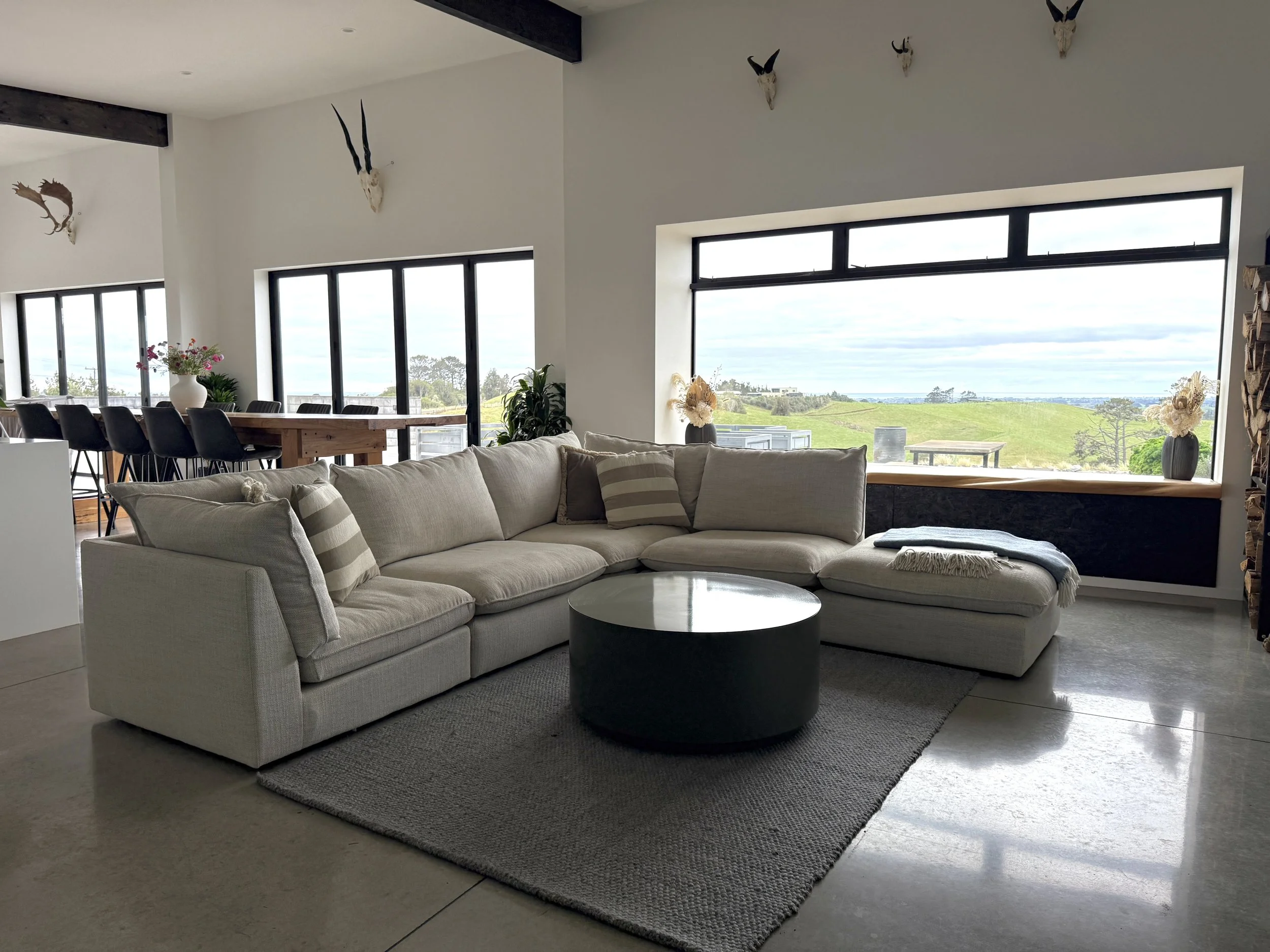Modern open-plan living room with a sectional sofa, round coffee table, dining area, large windows, and coastal countryside views