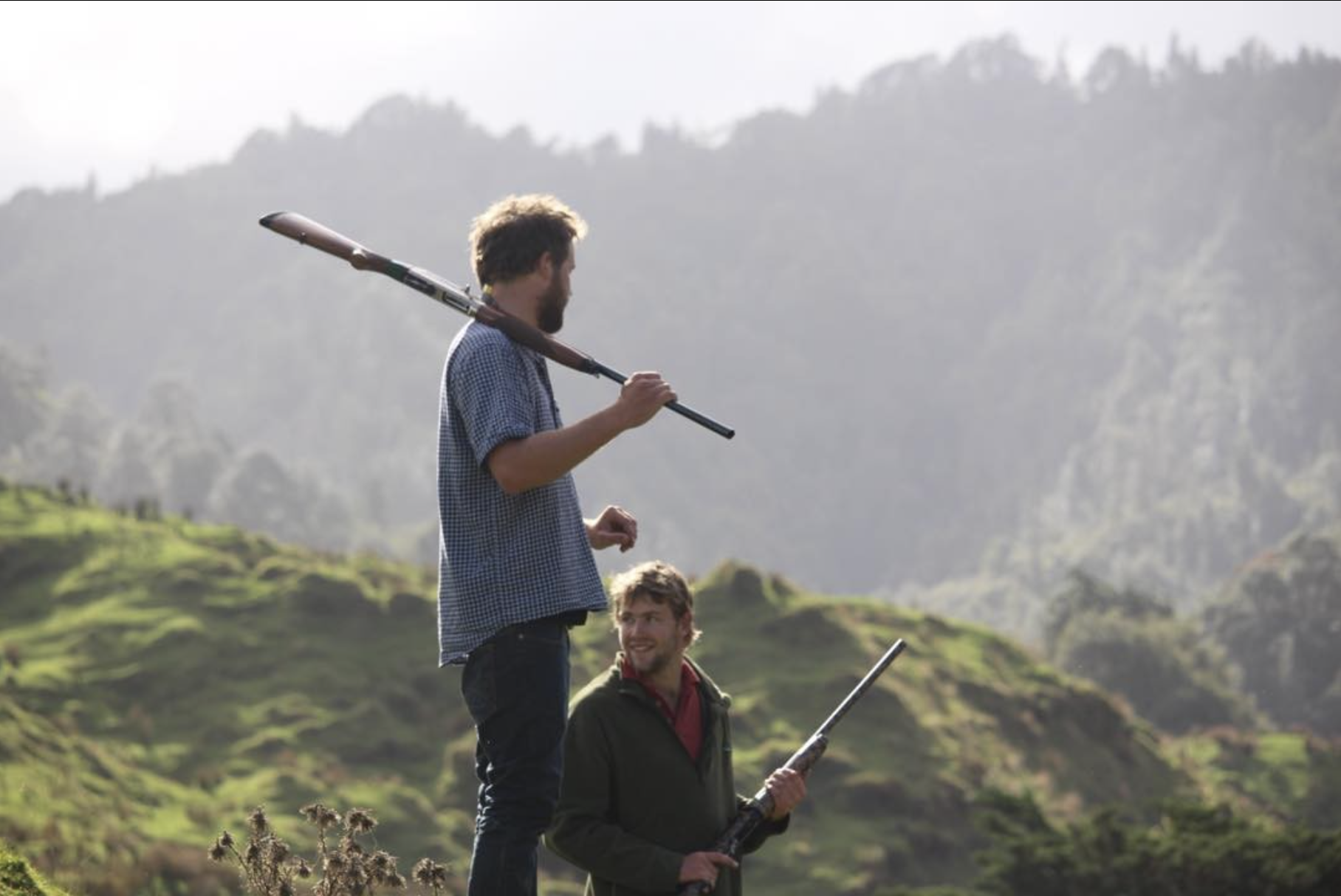 Two men carrying shotguns in a green, mountainous landscape