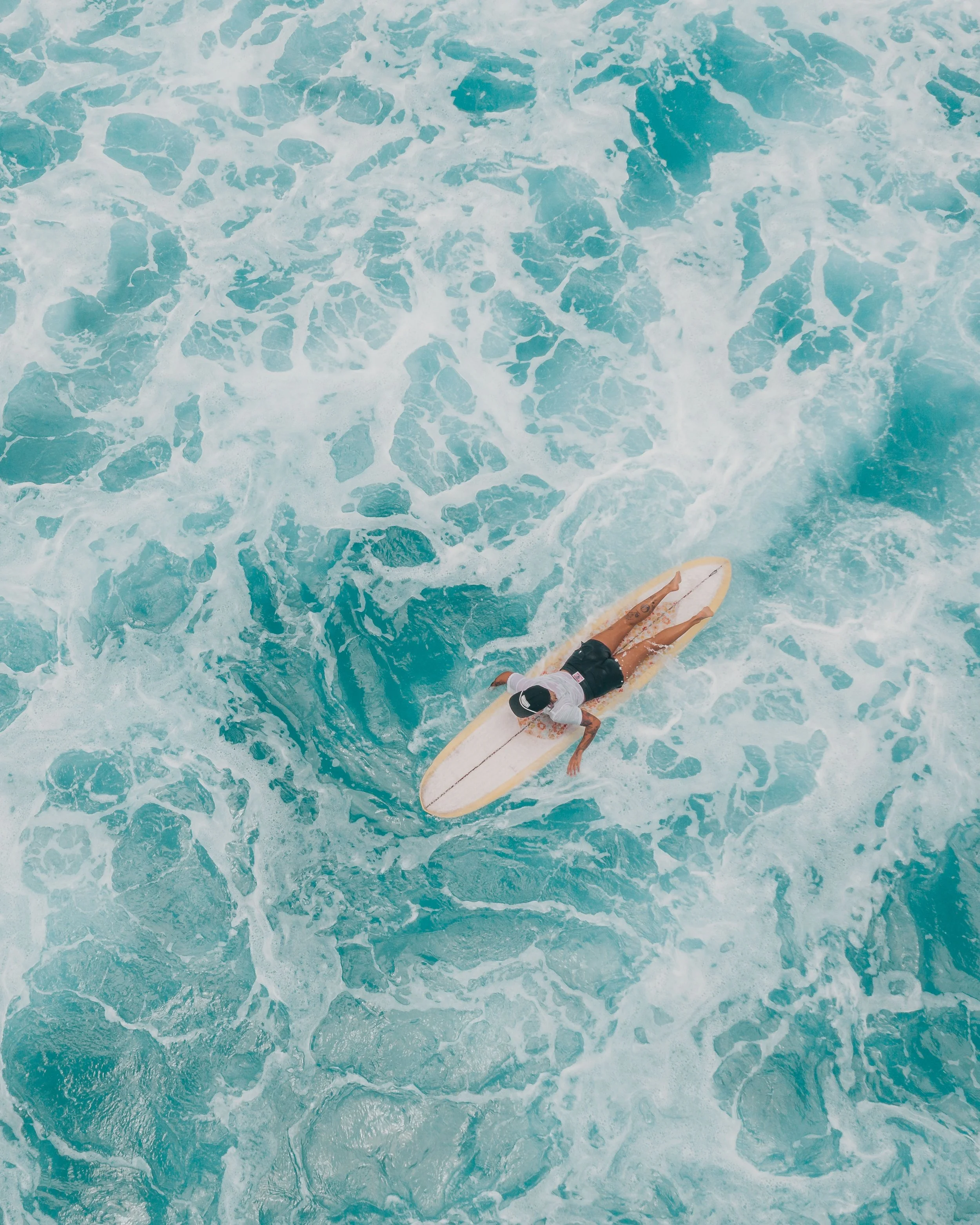 Aerial view of a surfer riding a longboard through turquoise ocean waves and white foam