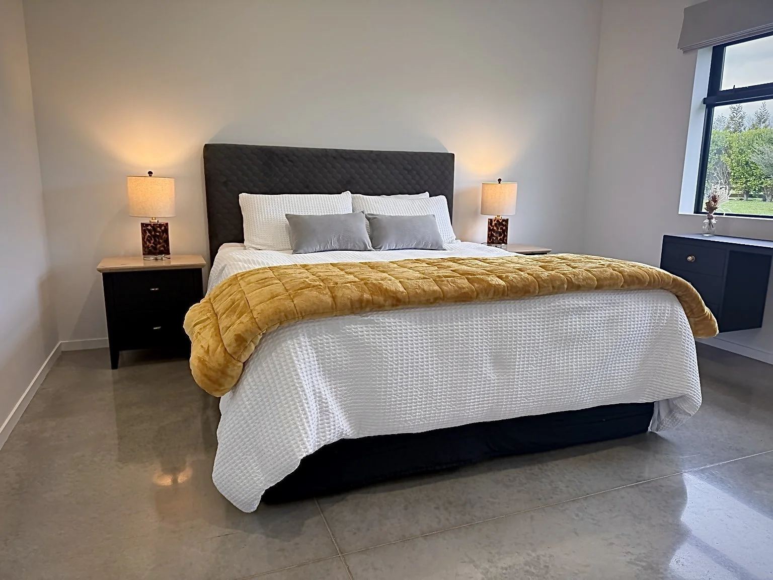 Modern bedroom with a king-size bed, white bedding, mustard-yellow throw blanket, bedside lamps, and a large window.