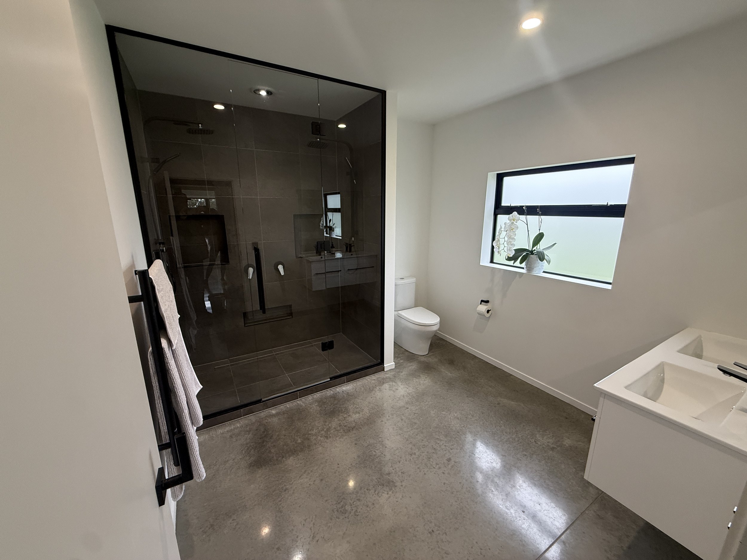 Modern bathroom with a glass-enclosed dark tile shower, freestanding bathtub, toilet, and orchid beneath a frosted window