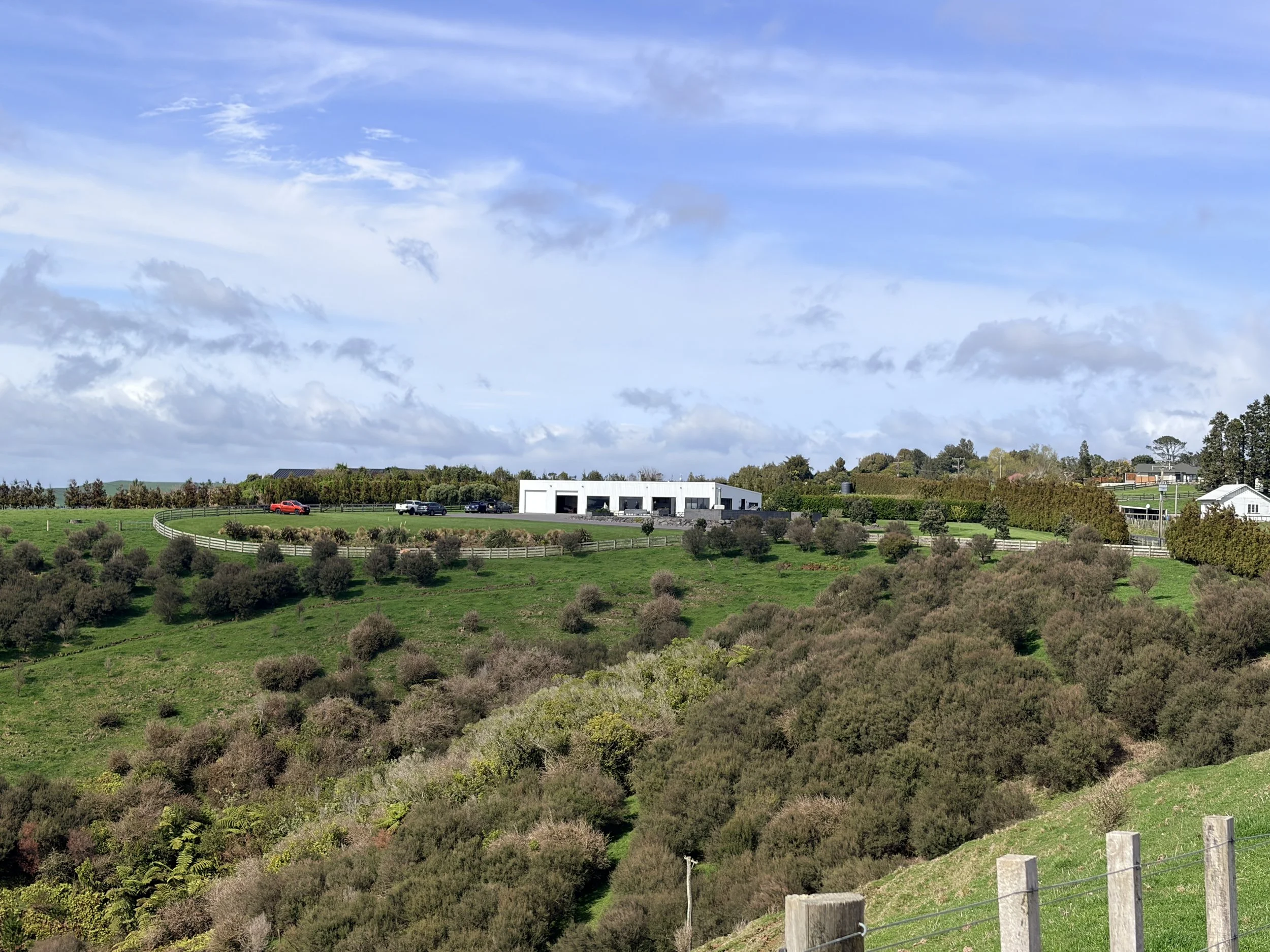 Rural green farmland with fenced fields, a modern white farm building, scattered homes, and a cloudy blue sky